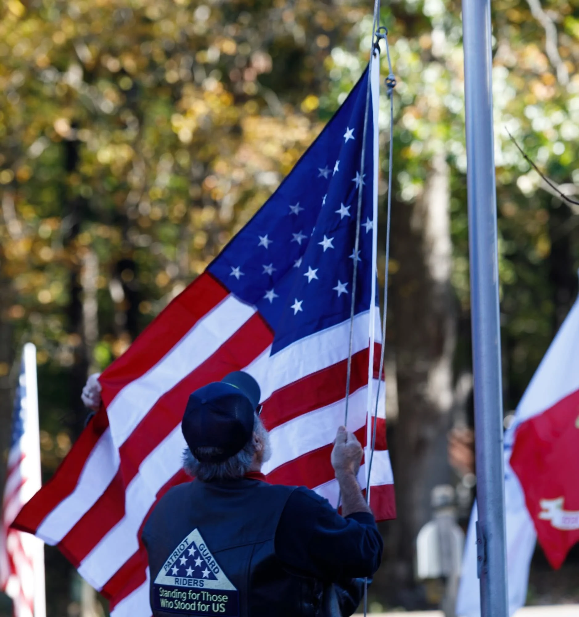 A person in a dark jacket and gloves raises a small American flag on a pole outdoors, with fall-colored trees in the background.