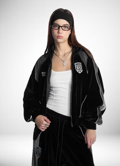 A young woman with long dark hair, wearing glasses, a black headband, a black jacket with a sports team logo, a white top, and black pants, standing against a plain white background.