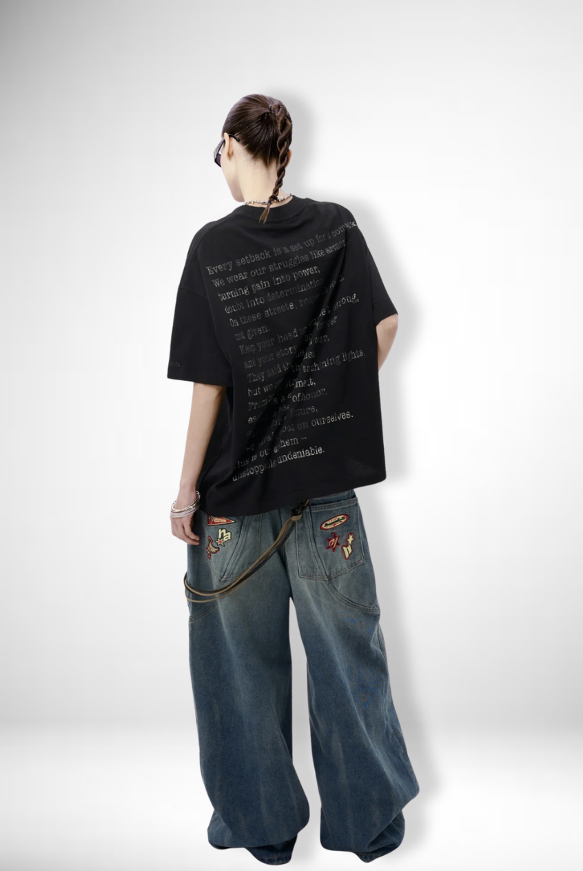 Person with long braided hair wearing sunglasses, oversized black T-shirt with printed text on the back, wide baggy jeans with patches, standing against a plain white background.