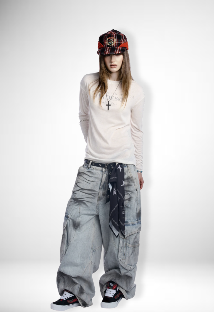 A young woman wearing a red and black plaid cap, white long-sleeve shirt, oversized baggy jeans, black sneakers with red accents, a black bandana tied around her waist, and a cross necklace standing against a plain white background.