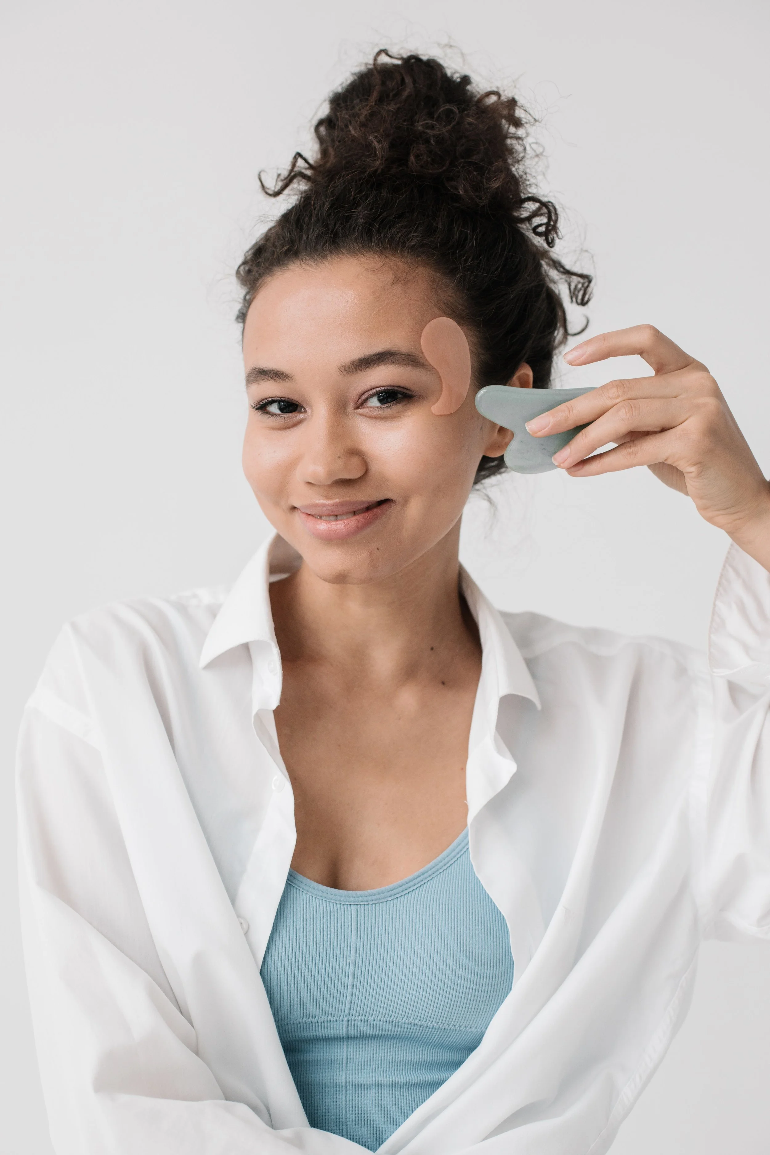 A woman in a white button-up shirt applying a skincare or beauty product to her face using a jade roller.