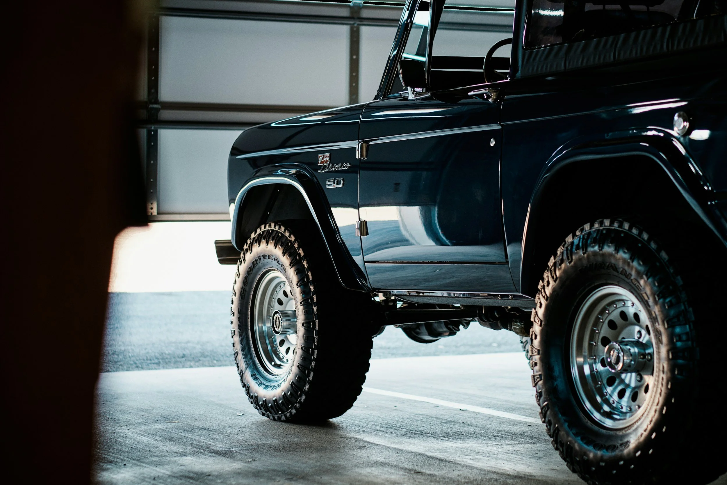 A close-up of a black Mercedes-Benz G-Class SUV parked inside a garage, showing part of the front and side of the vehicle, including the tires and side mirror.