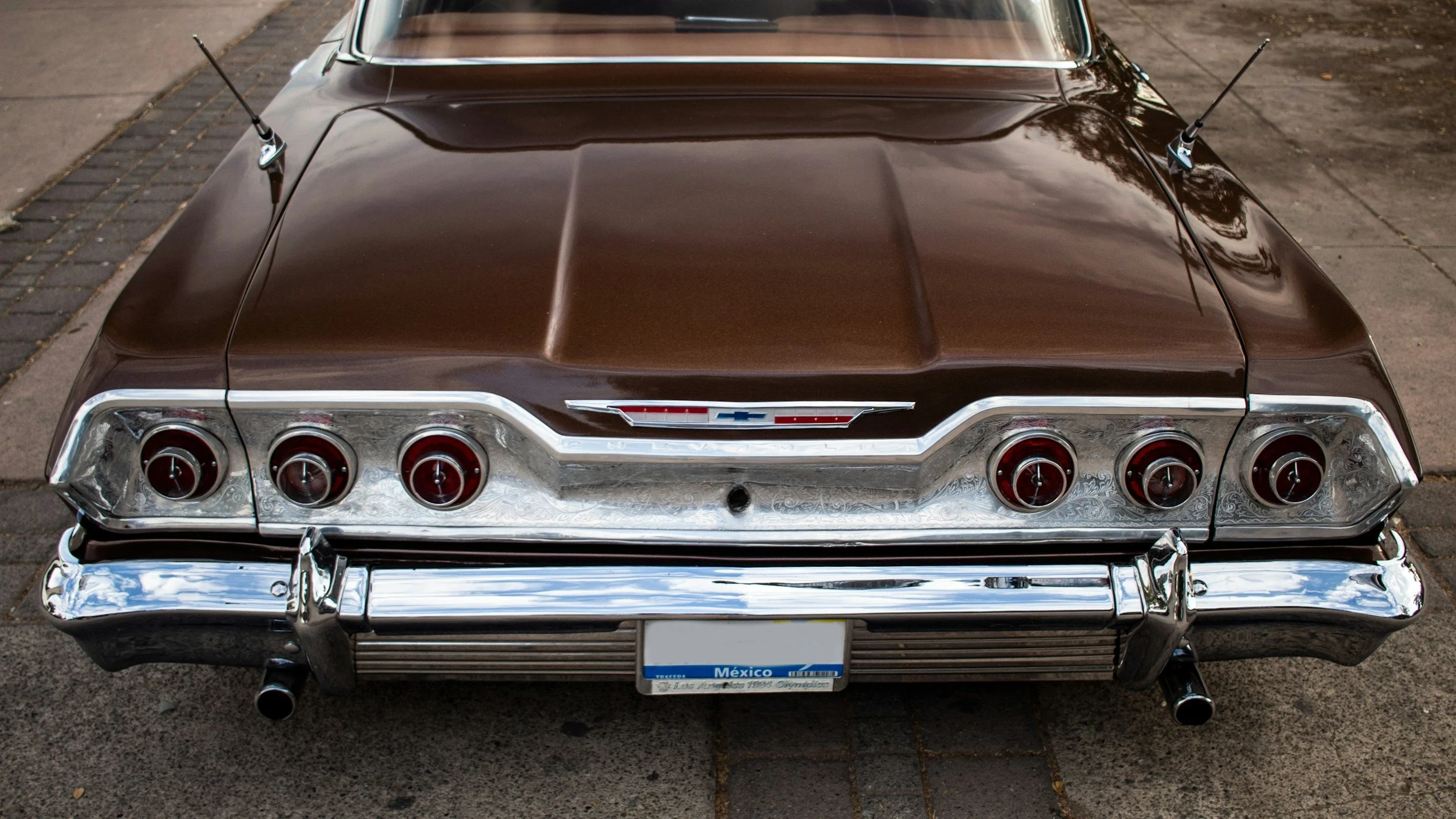 The back of a classic dark brown Chevrolet car with six round brake lights and a chrome bumper, parked on a sidewalk.