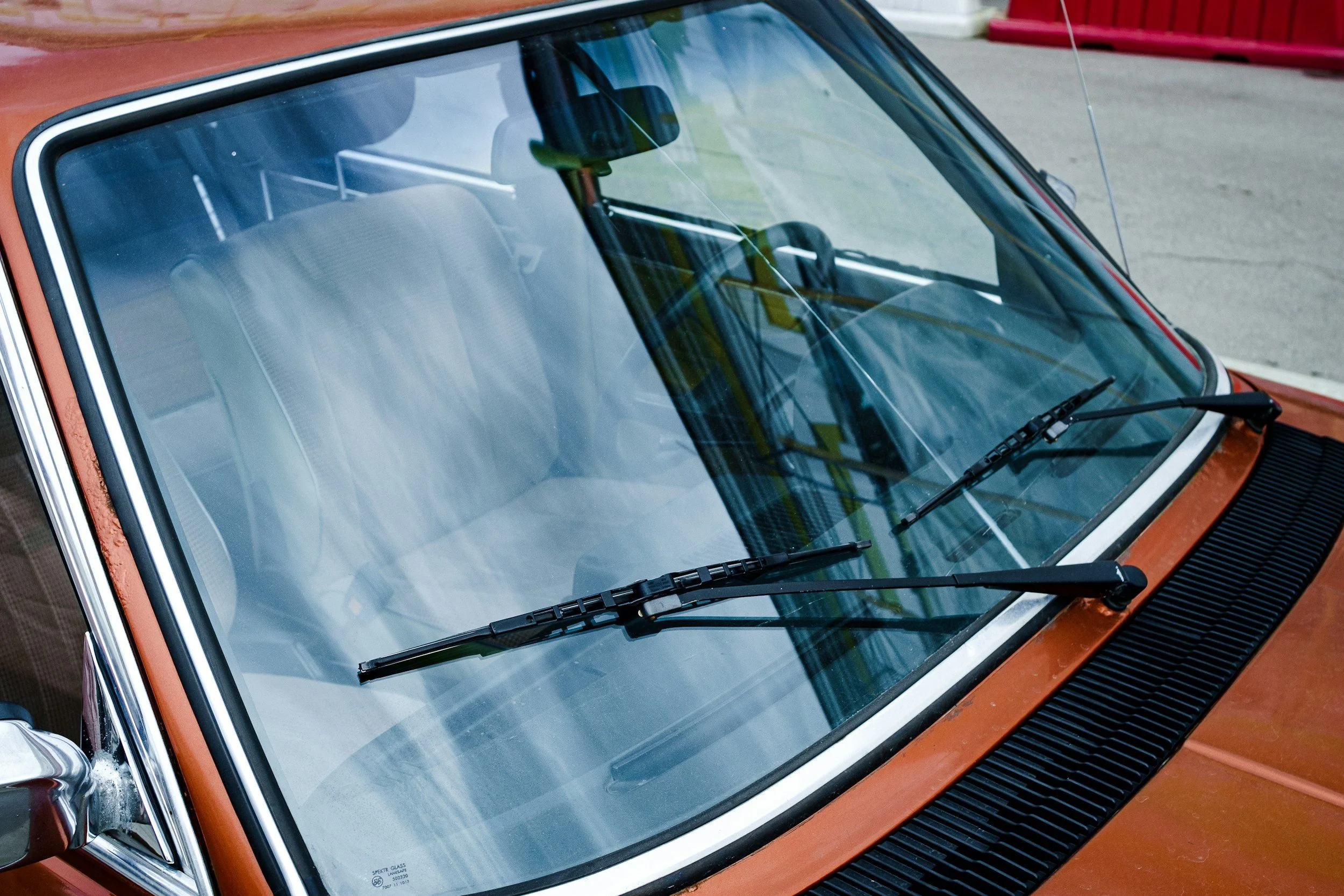Front windshield without rock chips of a vintage brown and black car with windshield wipers resting on the glass, parked on a paved surface with a red fence in the background.