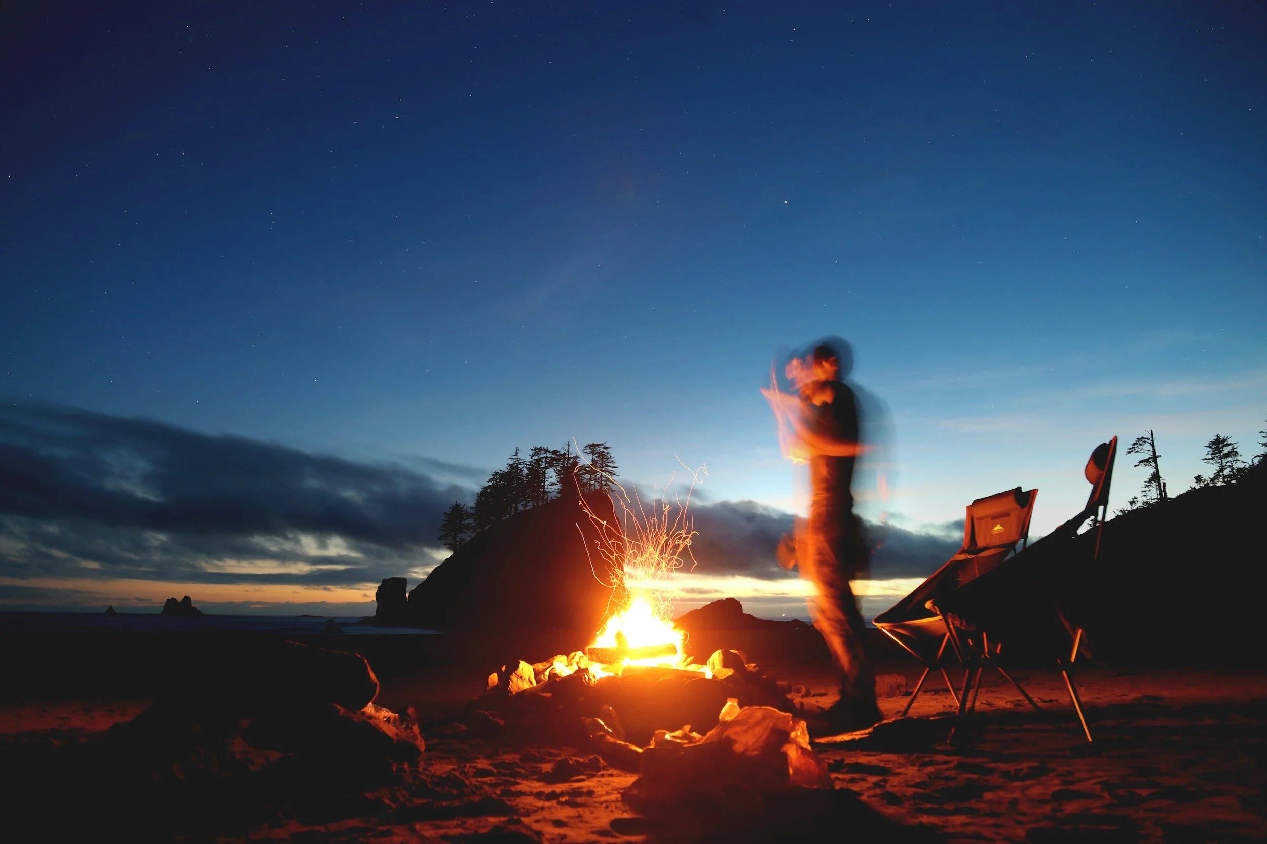 People sitting around a campfire on a beach at dusk, with chairs and trees in the background, and stars starting to appear in the sky.