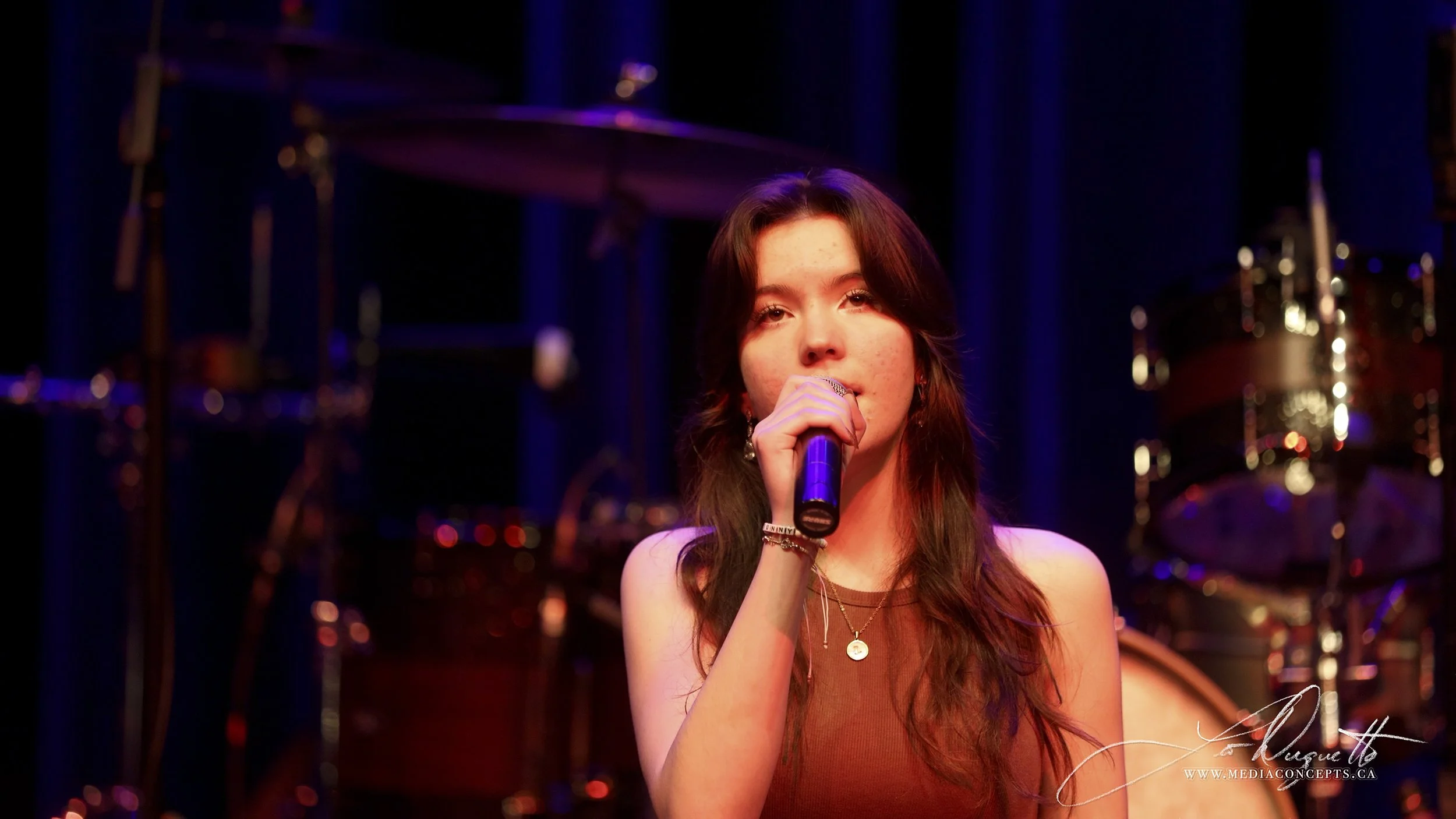 A young woman with long brown hair singing into a microphone on stage, with a drum set in the background, under stage lighting. Luna Fox.