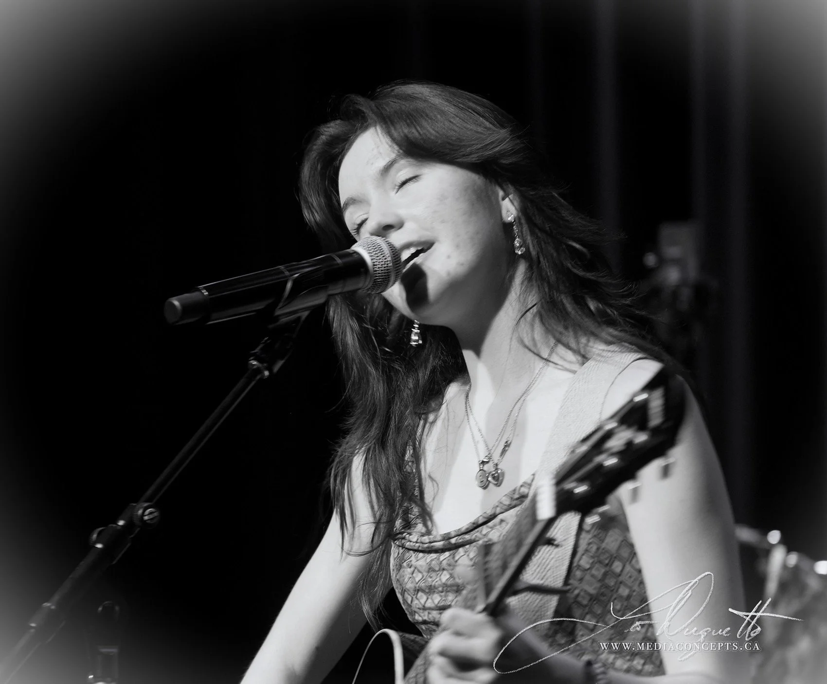 A woman singing into a microphone while playing an acoustic guitar, with her eyes closed, in a black and white photo. Luna Fox.