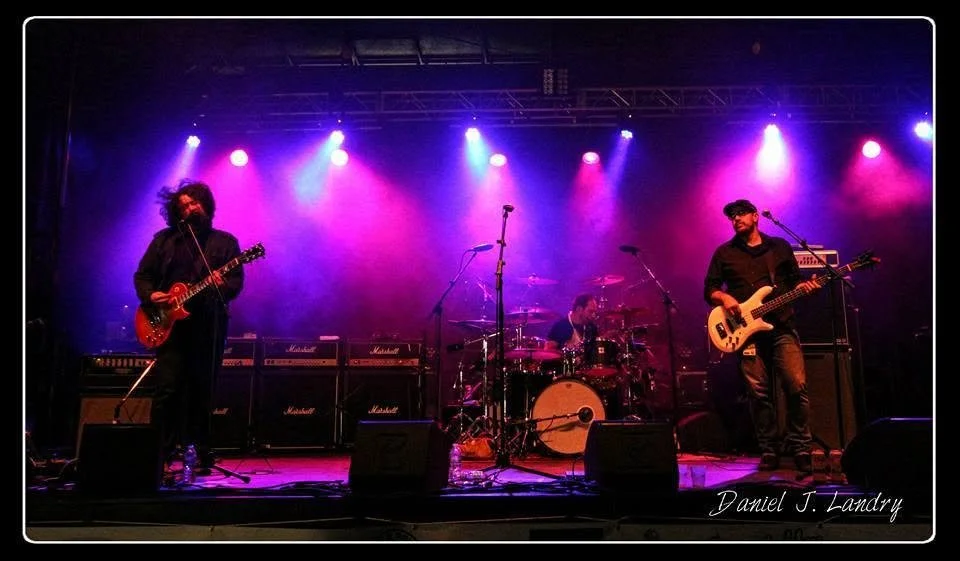 Toe Jam Tequila guitarist (Dayv Poulin), drummer (Neil Kaltiainen) and bassist (Guy Coutu) performing under purple and blue concert lights.