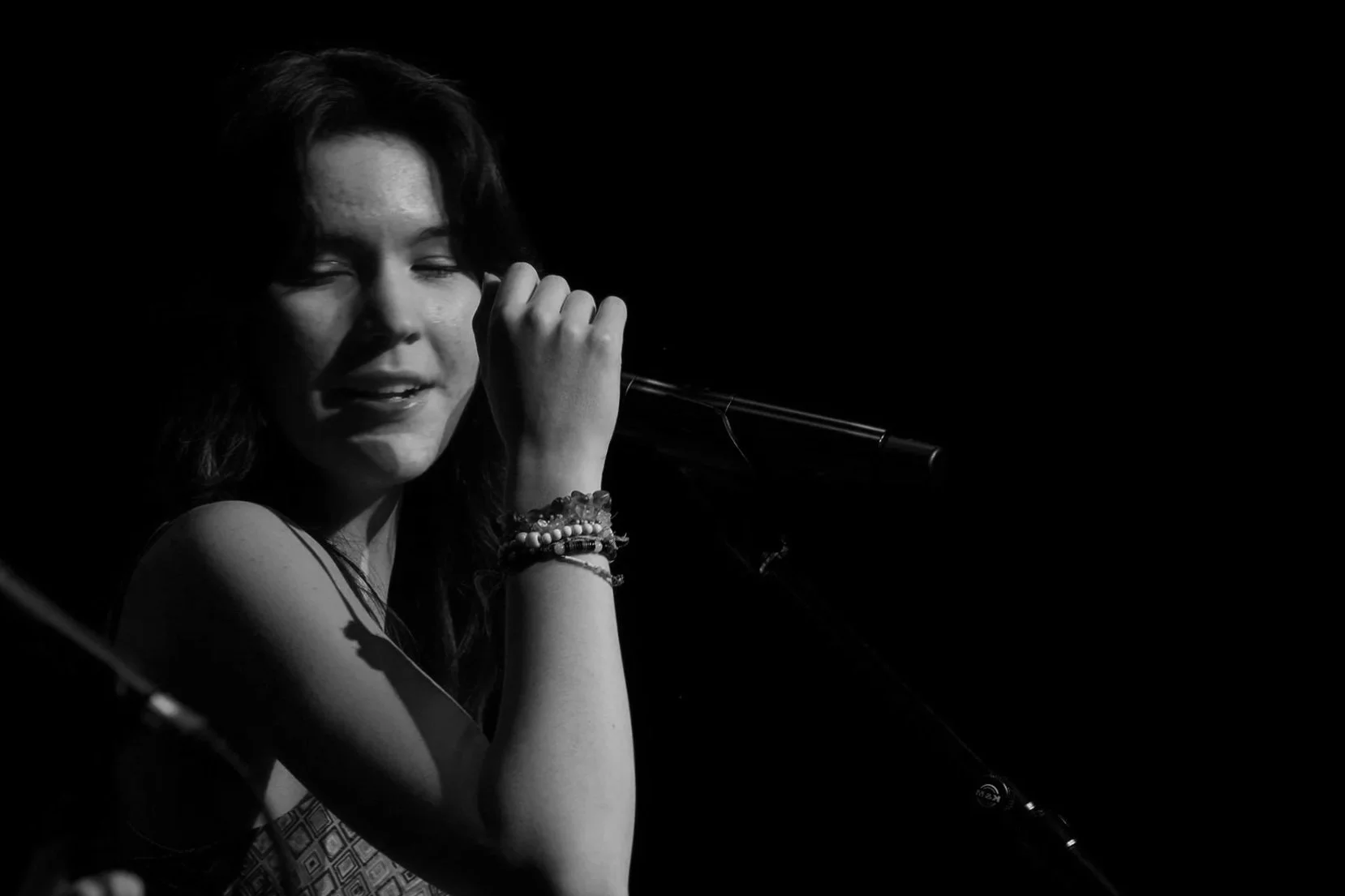 Black and white photo of a woman singing into a microphone with closed eyes, wearing bracelets, against a dark background. Luna Fox.