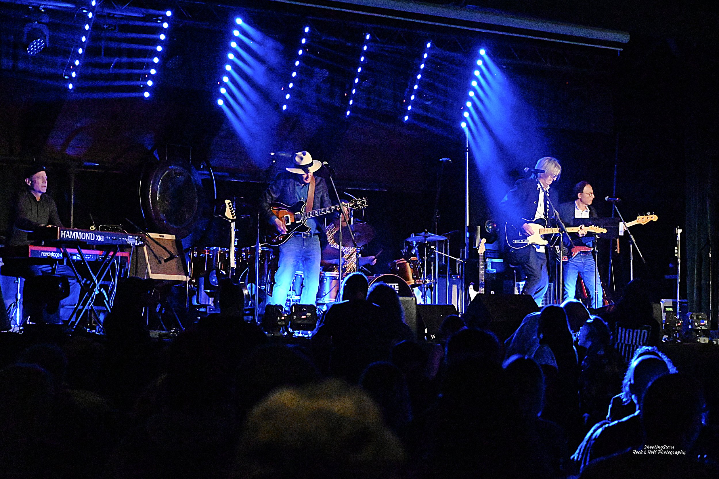 Tribute - Canada's premiere Tom Petty experience, live music performance with a band on stage playing guitars, keyboard, and drums, under blue stage lights, with an audience in front.