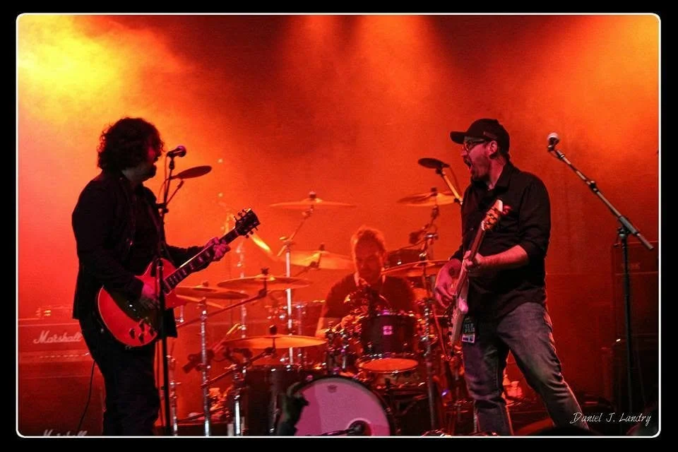 Toe Jam Tequila guitarist (Dayv Poulin)and bassist (Guy Coutu) facing each other onstage as the drummer (Neil Kaltiainen)  performs behind them under red and orange lighting.
