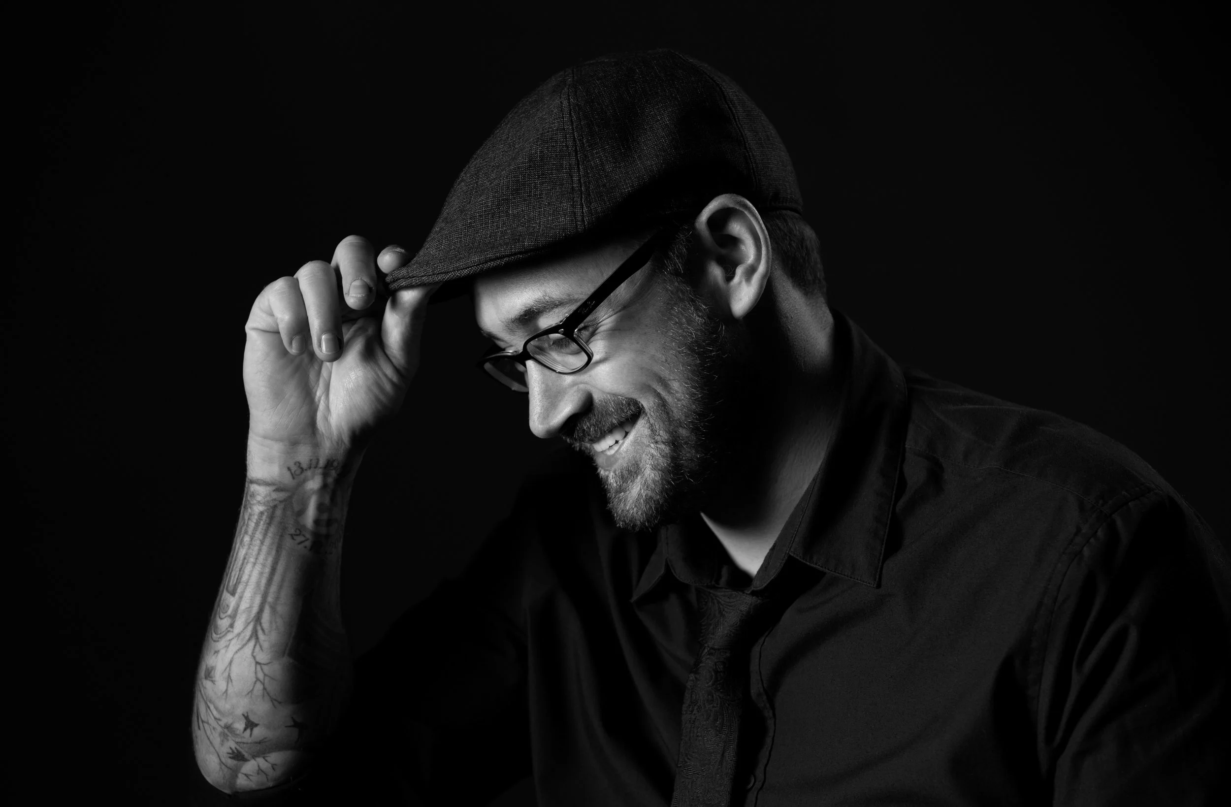 Wax Cylinder Music. A man in a cap and glasses is smiling and holding the brim of his cap with his right hand, facing downward, in a black-and-white portrait.