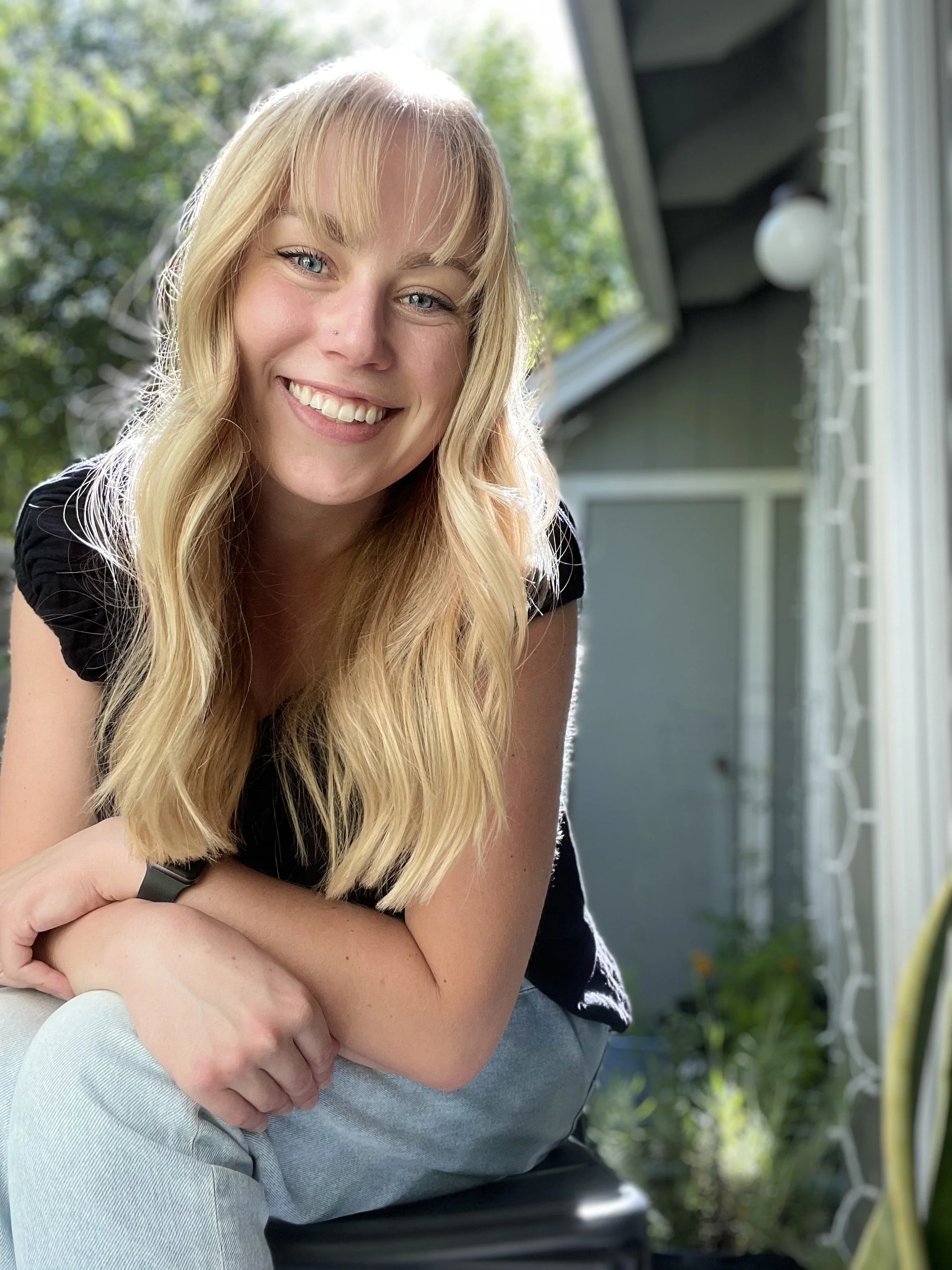 A woman with long blonde hair smiling outdoors near a house, with a background of green trees and a gray house.