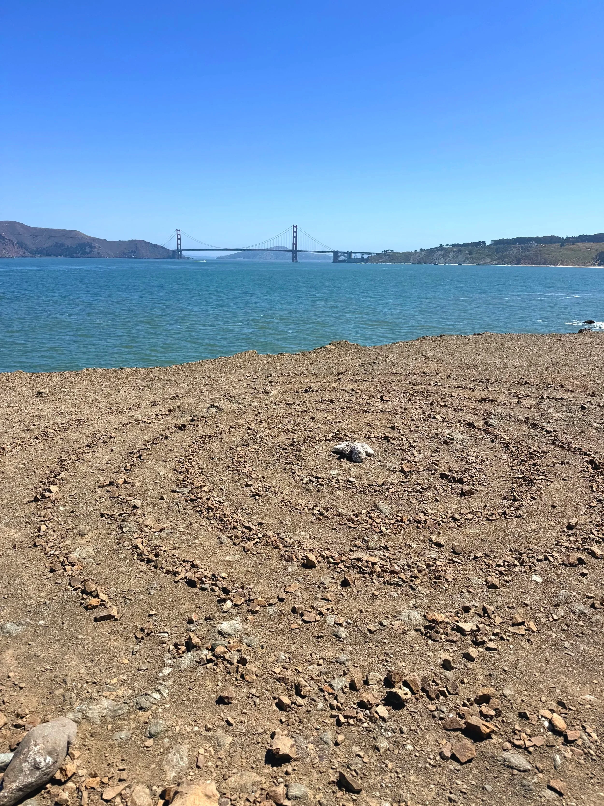 View of the Golden Gate Bridge and labyrinth in San Francisco, with water in the foreground and hills on the sides, under a clear blue sky.