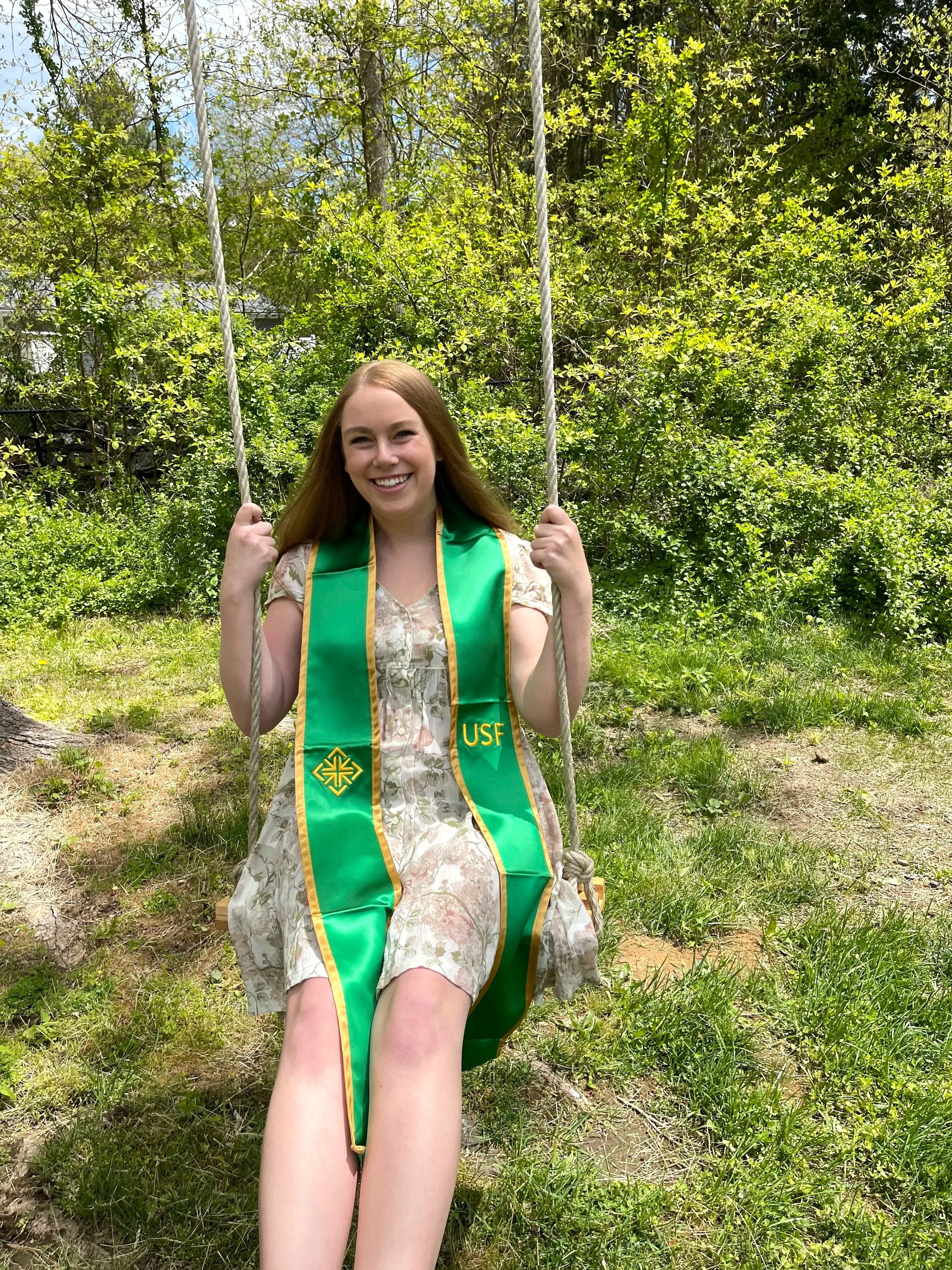 A young woman with long blonde hair sitting on a swing in a garden, wearing a floral dress and a green graduation stole with yellow trim that has the letters "USF" on it. She is smiling with a background of lush green trees and grass.