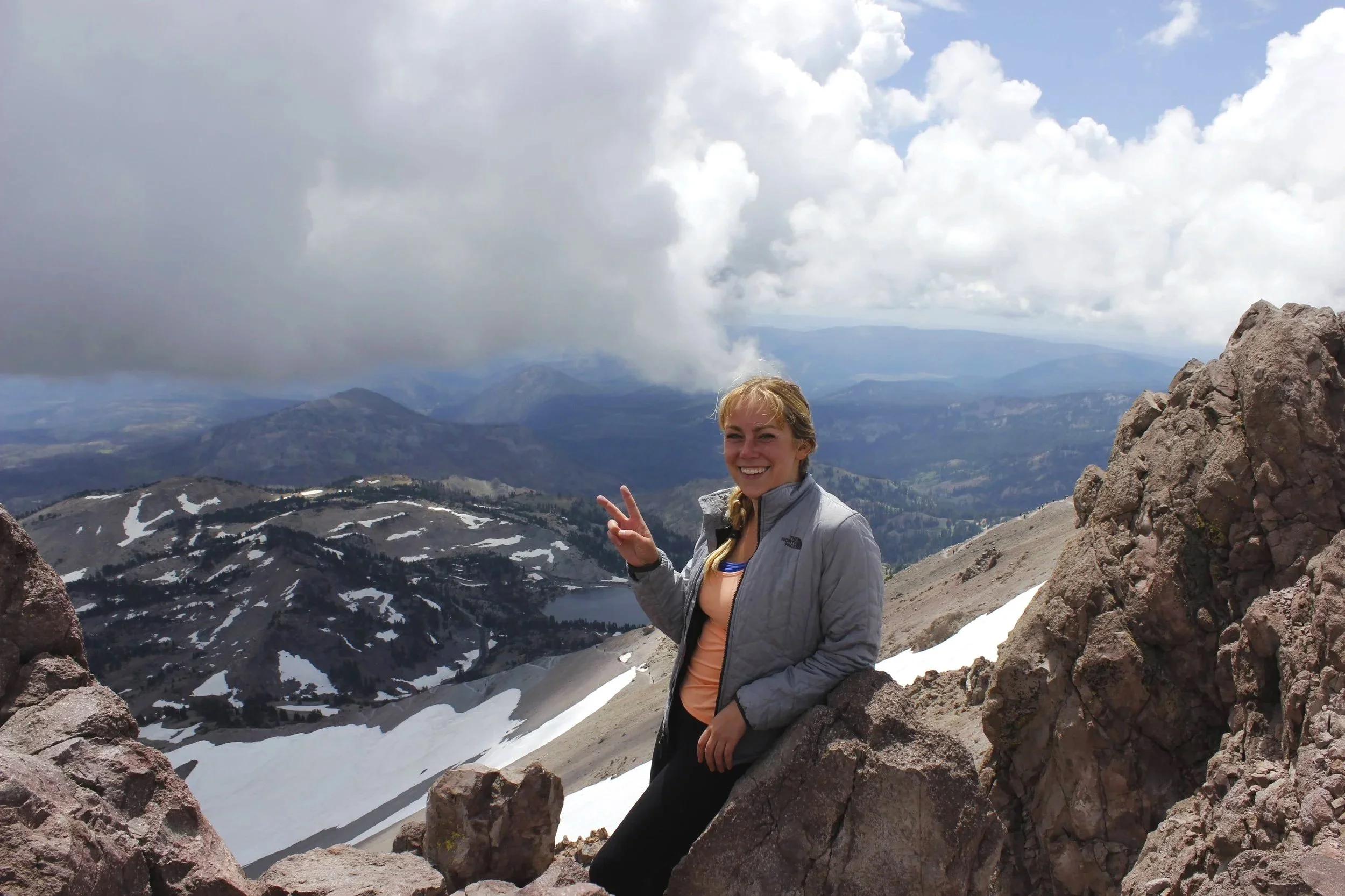 A woman with blonde hair smiling and making a peace sign with her fingers, standing on a mountain with snow patches and rocky terrain, with a cloudy sky and mountain landscape in the background.