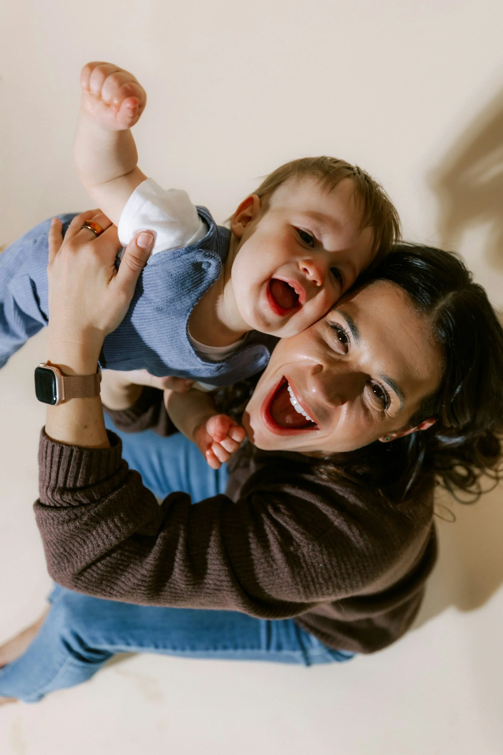 A woman and a young boy lying on the floor, smiling and playing. The woman has dark hair and is wearing a brown striped sweater with a watch on her wrist. The boy has light brown hair, is wearing a gray and white shirt, and has his arm raised in a fist.