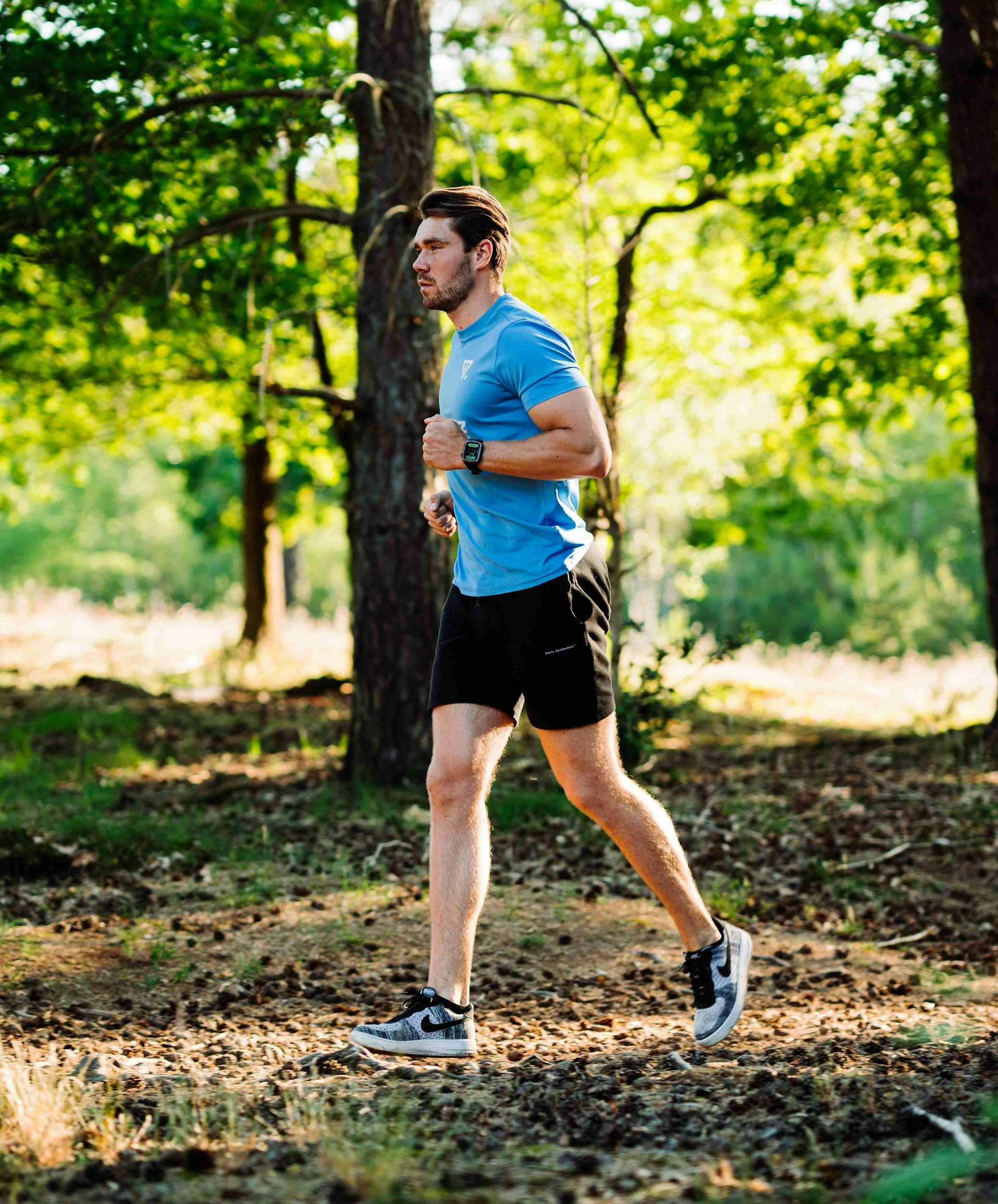A man running outdoors in a wooded area wearing a blue t-shirt, black shorts, sneakers, and a smartwatch.