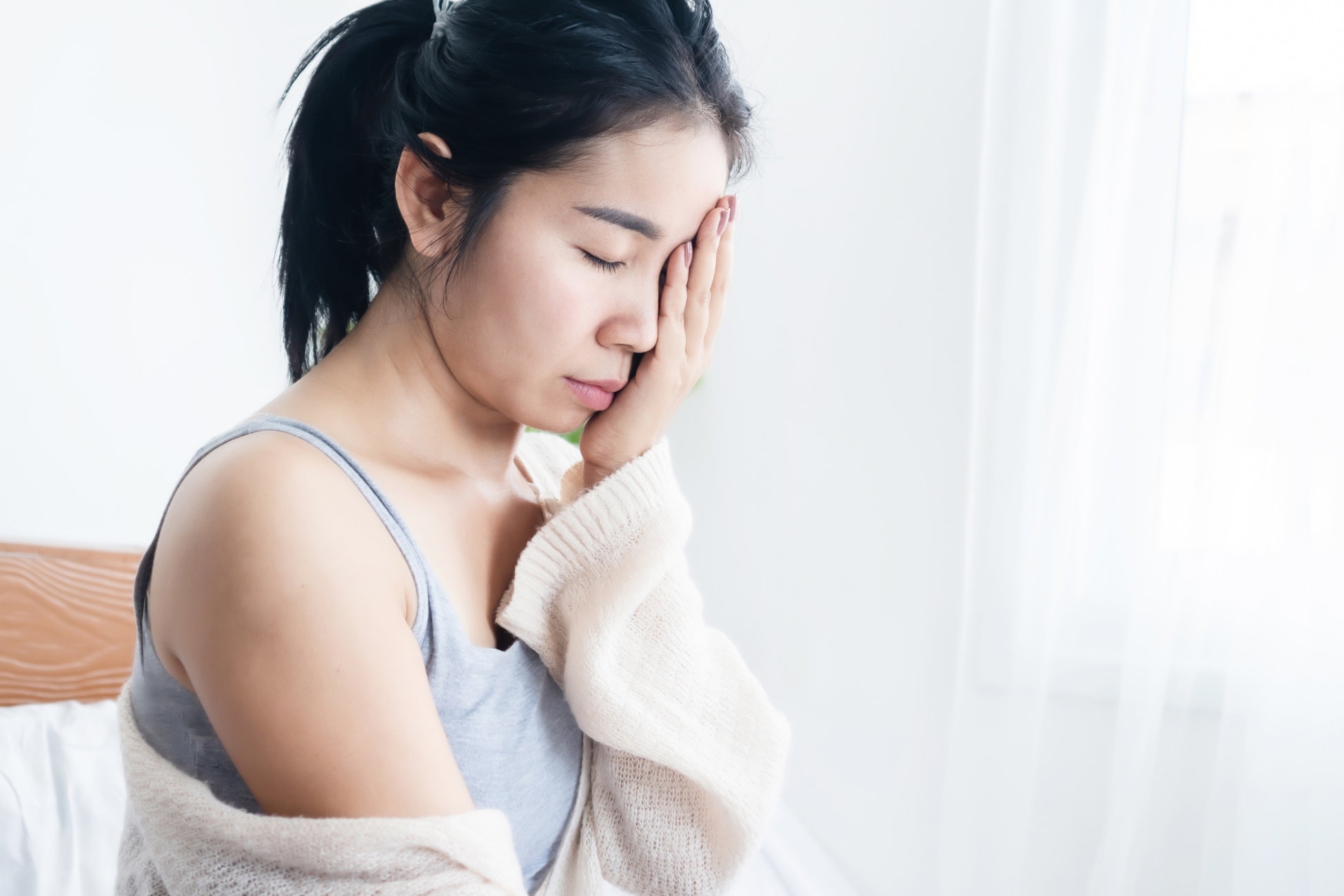A woman with long black hair sitting on a bed, holding her head with her hand, appearing to be in distress or pain.