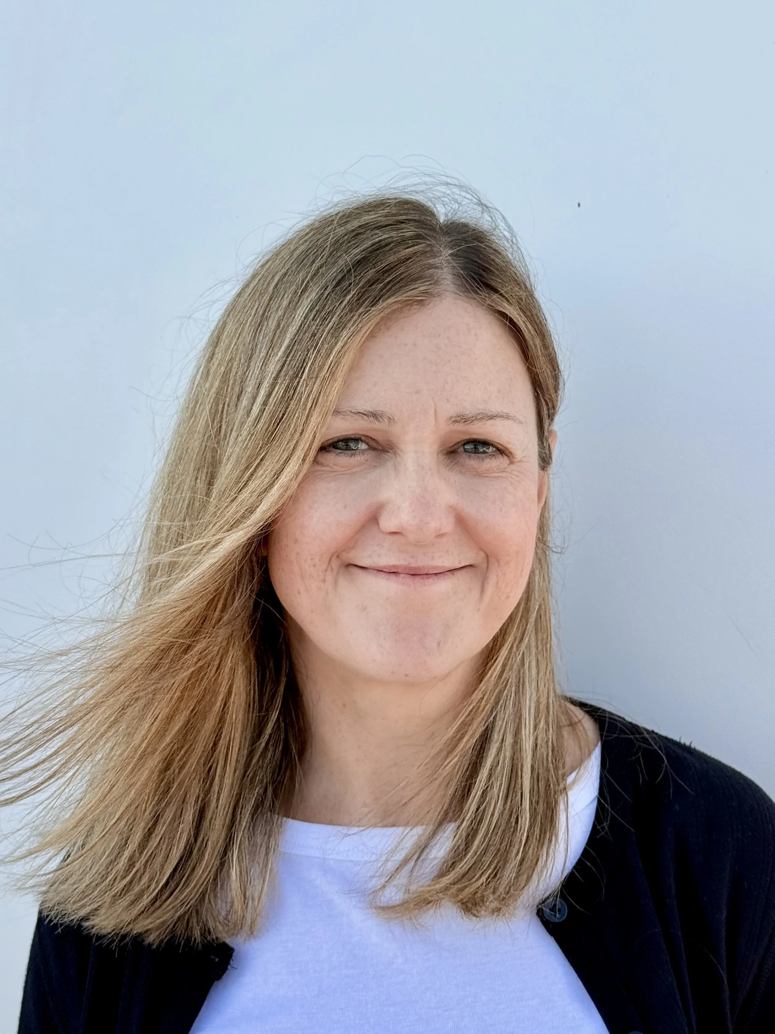 Close-up of a smiling woman with long blonde hair, wearing a white shirt and a black cardigan, against a light blue background.