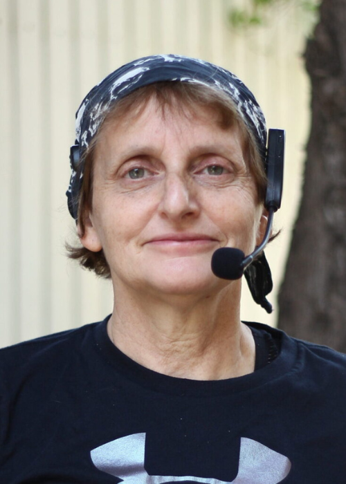 A woman with short light brown hair wearing a black shirt and a headset with a microphone, standing outdoors in front of a tree.
