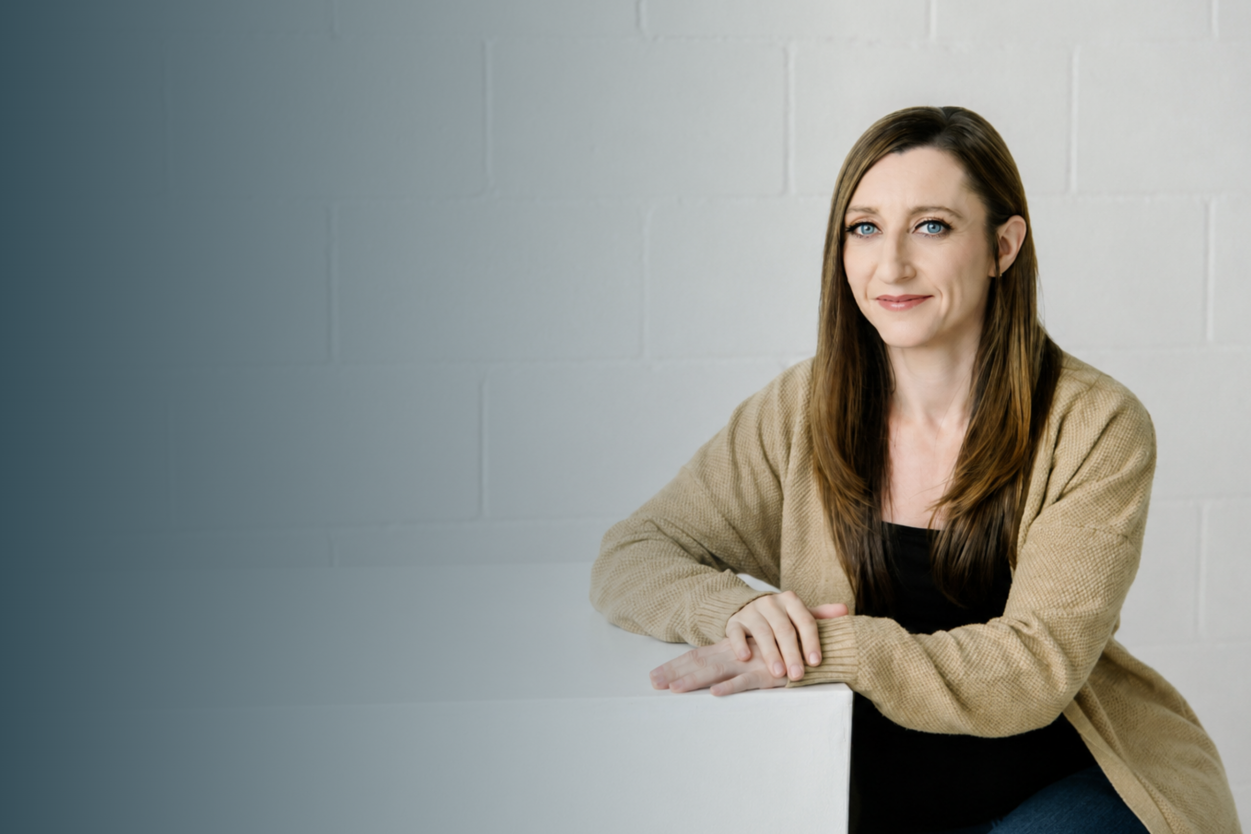 A woman with brown hair, blue eyes, and light skin sitting at a white table, smiling, wearing a beige jacket and black top, in front of a gray brick wall.