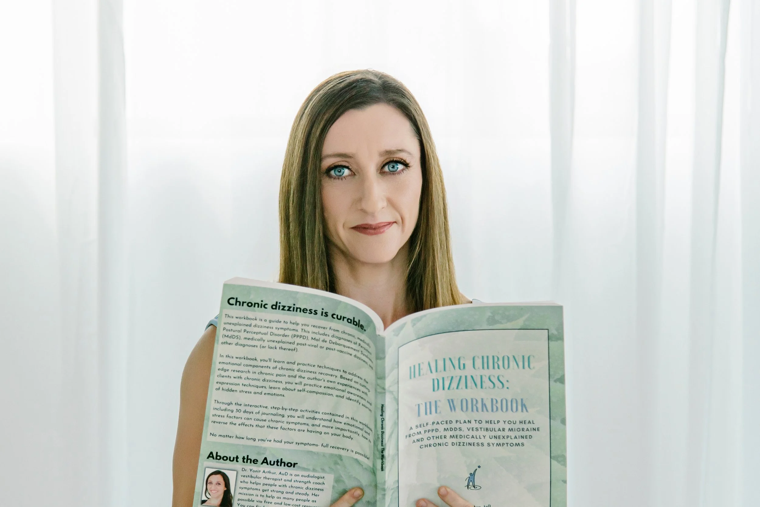 A woman with long brown hair and blue eyes reading a pamphlet titled "Healing Chronic Dizziness: The Workbook" in front of sheer white curtains.