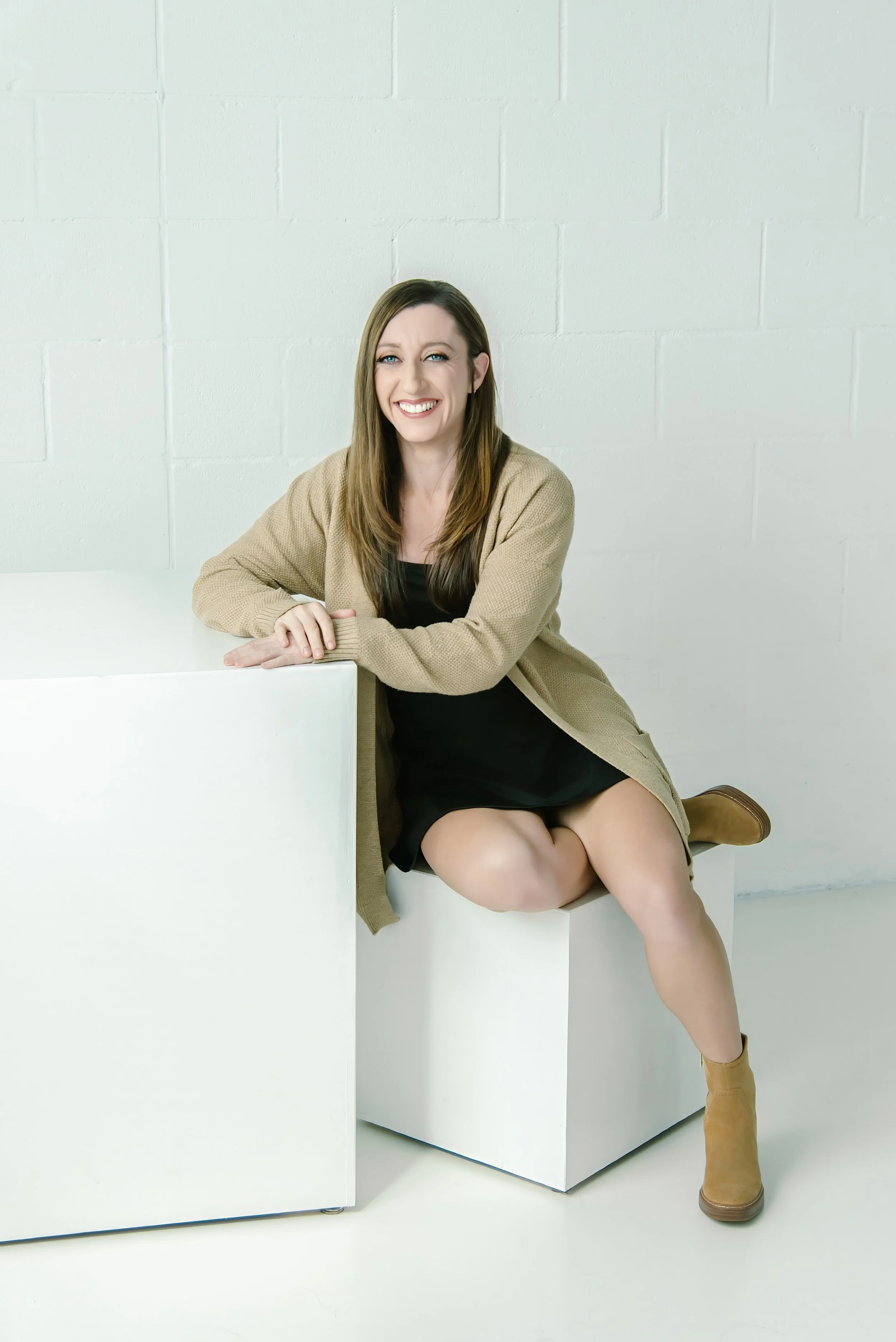 A woman sitting on a white cube, smiling, wearing a black dress, tan cardigan, and tan boots, against a white brick wall background.