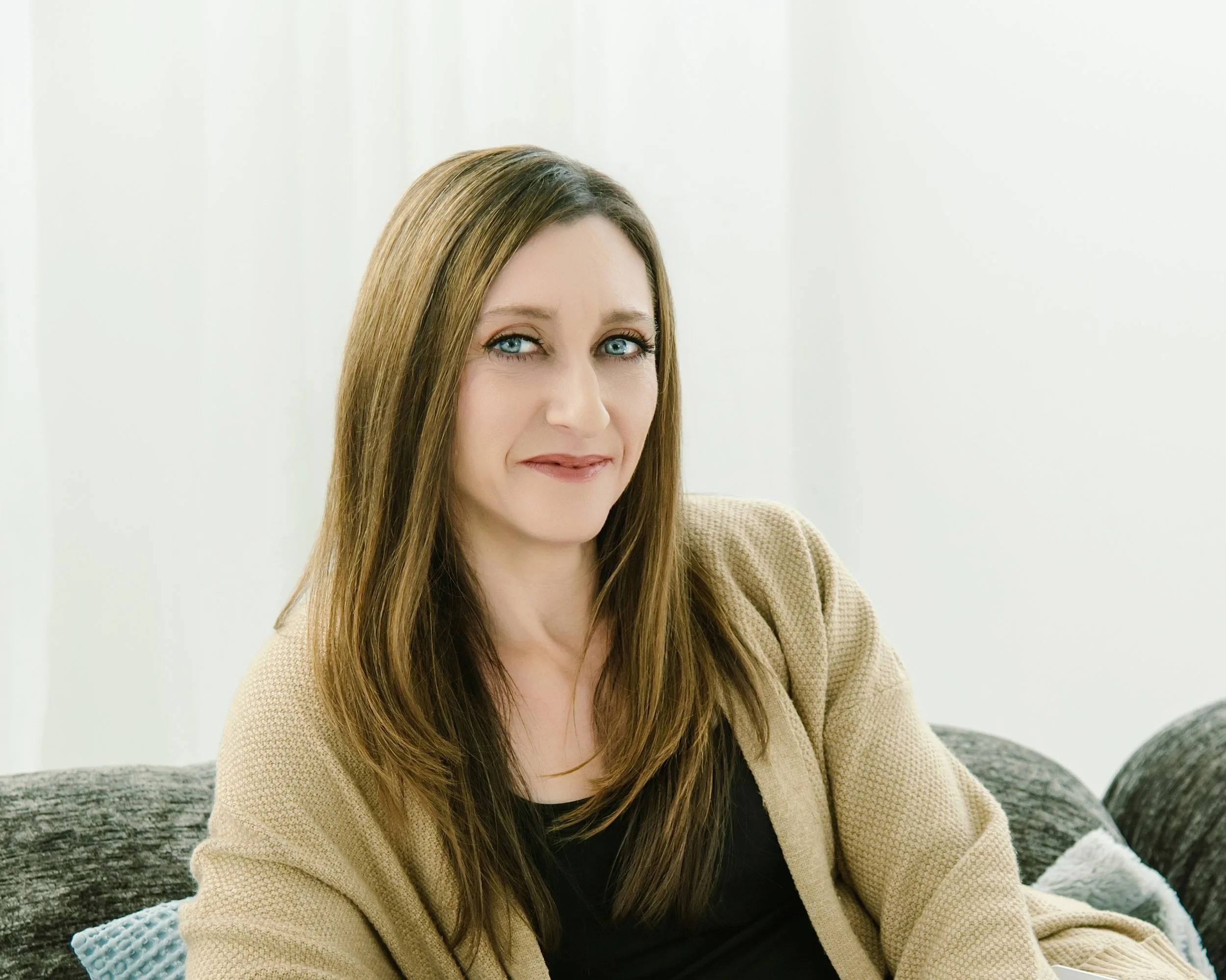 A woman with long brown hair and blue eyes sitting on a couch, smiling, wearing a black top and a beige cardigan.