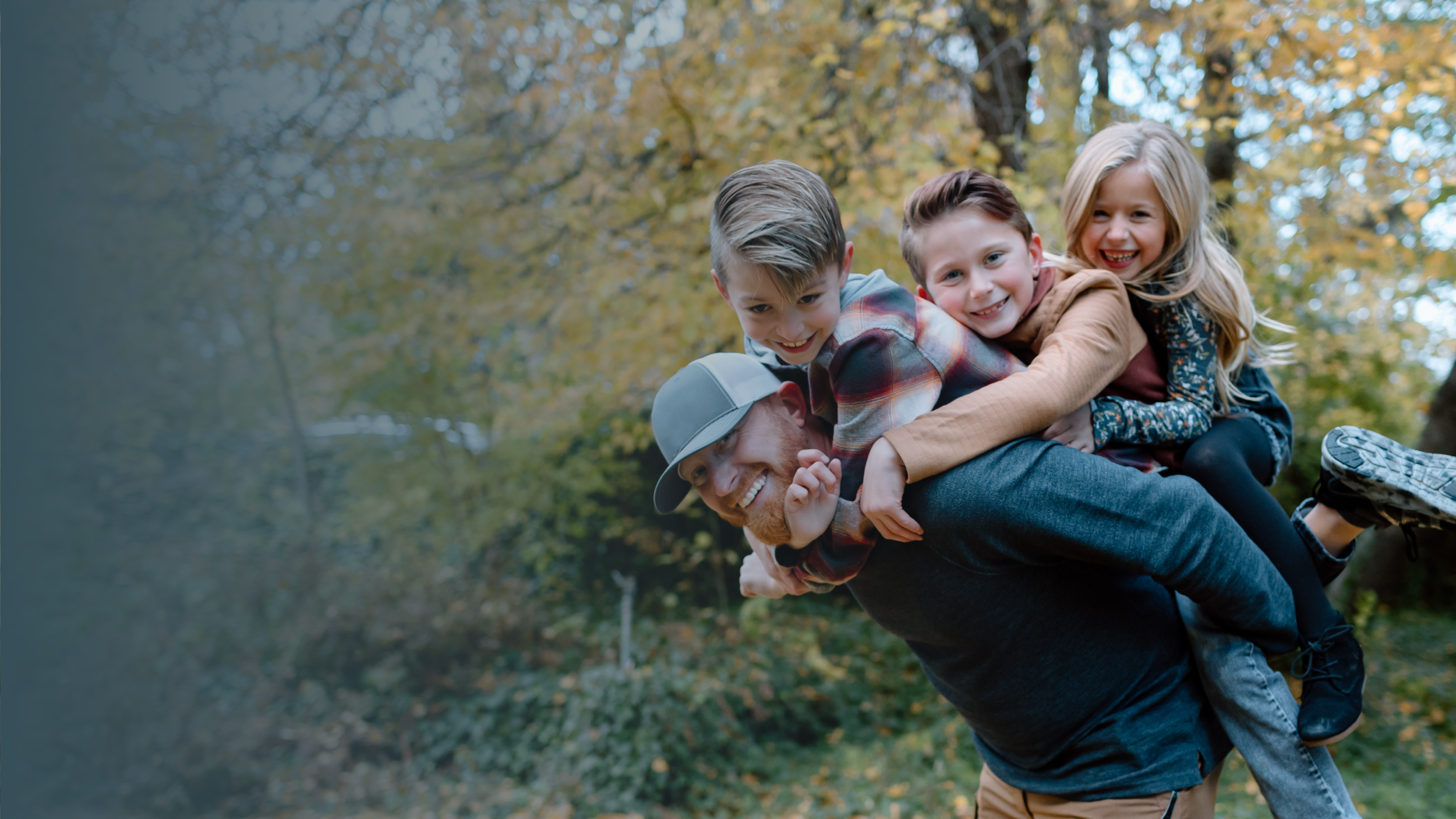 Four children and one adult outdoors, with trees and autumn foliage in the background. The adult is giving a piggyback ride with three children on his back, all smiling and enjoying themselves.