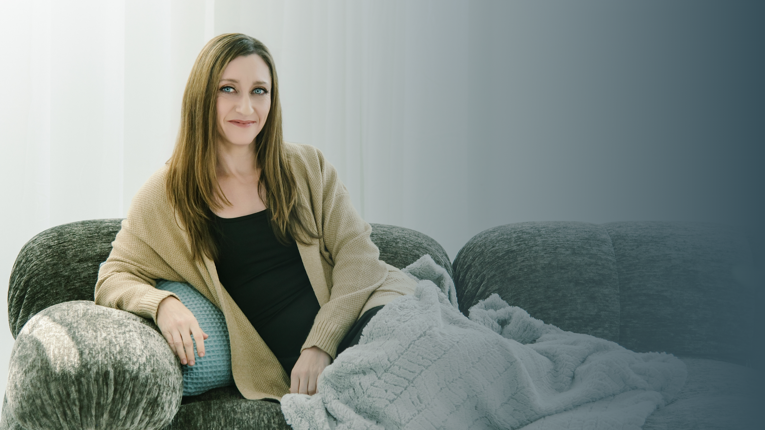 A woman with long brown hair and blue eyes sitting on a dark gray sofa, holding a gray blanket, wearing a black top and beige cardigan, in a well-lit room with white vertical blinds.