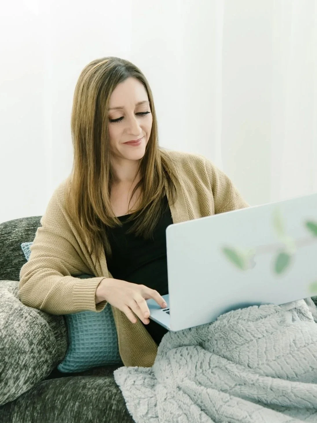 A woman with brown hair working on a laptop while sitting on a sofa with a blanket.