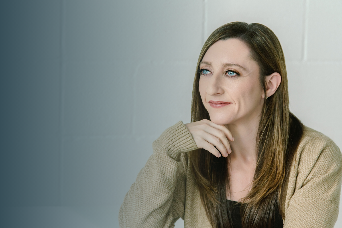 A woman with long brown hair and blue eyes, wearing a beige sweater, smiling gently, and resting her chin on her hand against a light-colored wall.