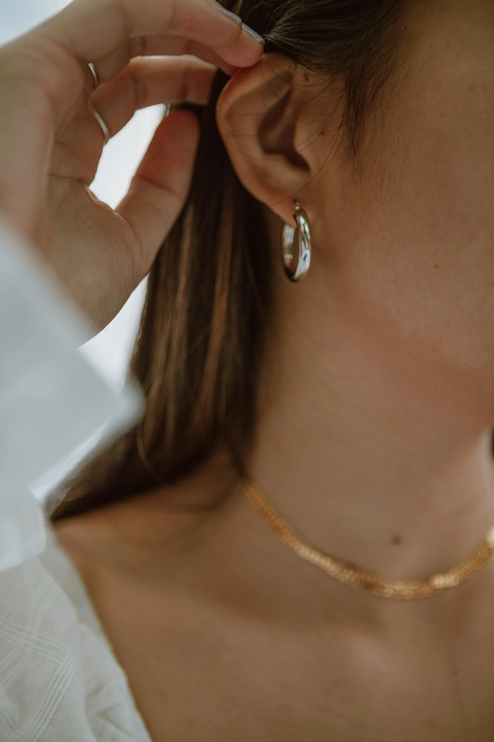 A person adjusting their earrings with their hand, showing part of their ear, neck, and a gold chain necklace. The person has brown hair and skin.