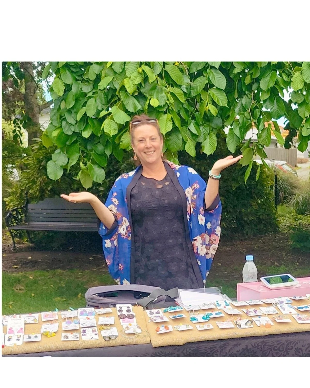 A woman standing outdoors behind a table displaying jewelry and accessories, smiling with arms raised. The table has multiple jewelry pieces, and there is a water bottle and a tablet on it. She is wearing a black dress with a floral kimono jacket.