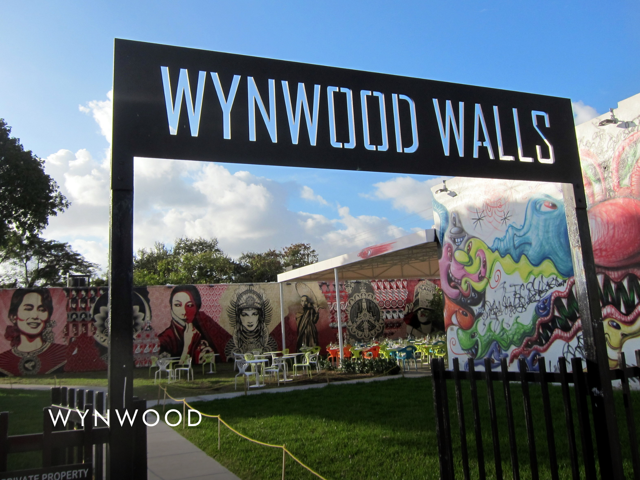 Sign with 'WYNDWOOD WALLS' in front of a mural with portraits and graffiti art, outdoor area with tables and chairs, partly cloudy sky.