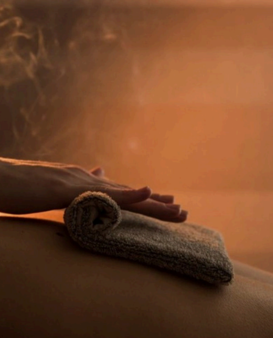 Person's hand gently pressing a rolled towel on their stomach, with steam rising in a warm, softly lit room.