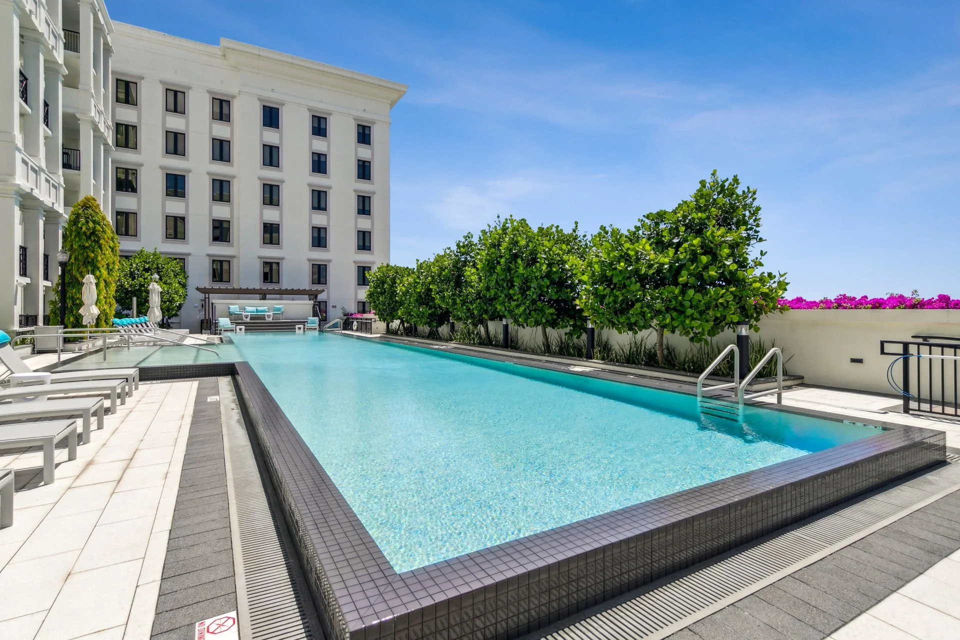 Image of Life Time Living at Gables Station, Coral Gables, showing an empty outdoor swimming pool on a sunny day with lounge chairs, a white building in the background, and green trees along the poolside, under a clear blue sky.