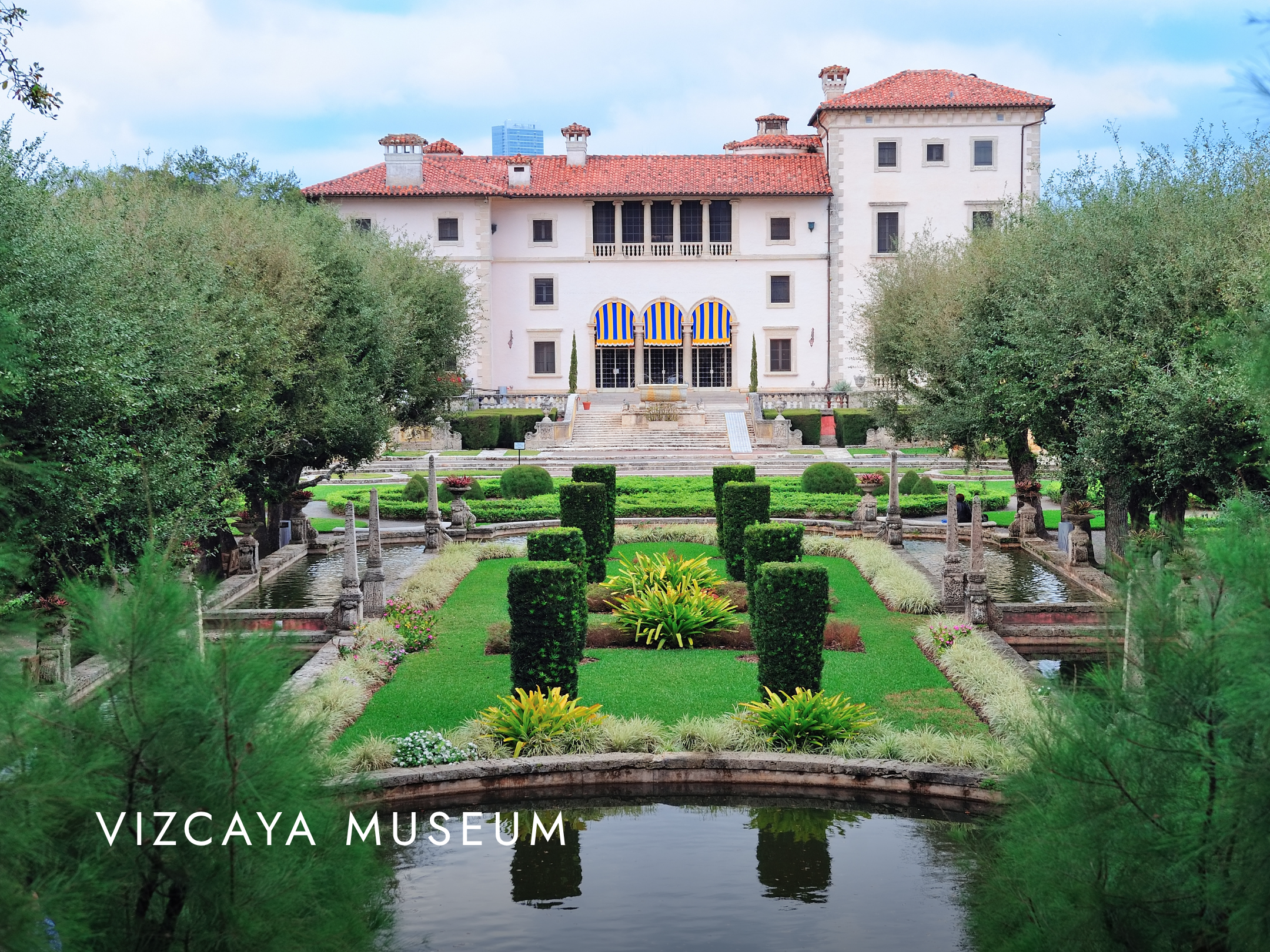 Viscaya Museum building with a red tile roof and multiple chimneys, surrounded by lush greenery and gardens, with the words "VIZCAYA MUSEUM" at the bottom.