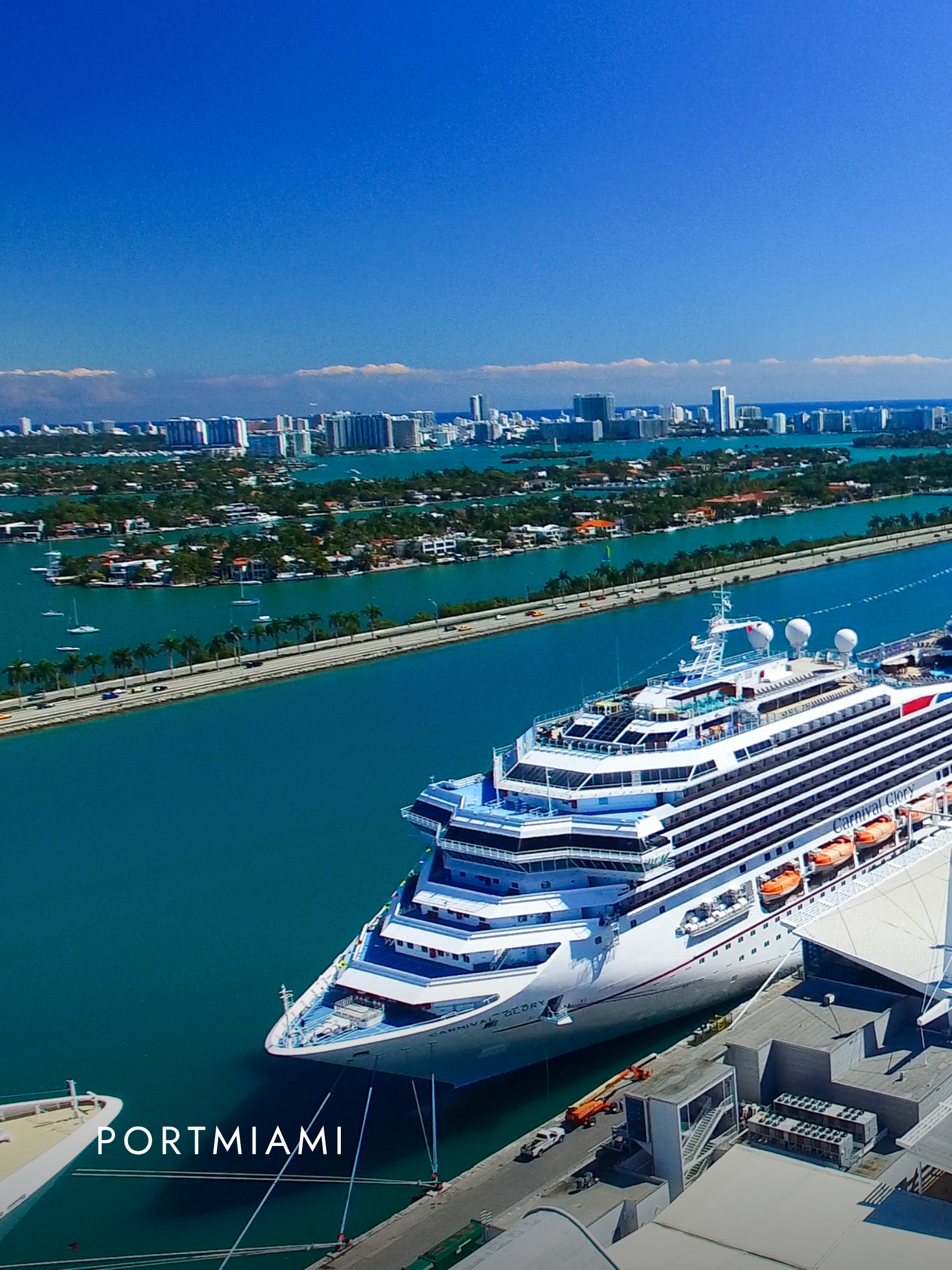 A large white cruise ship docked at a port in PortMiami, with city high-rise buildings and a clear blue sky in the background.