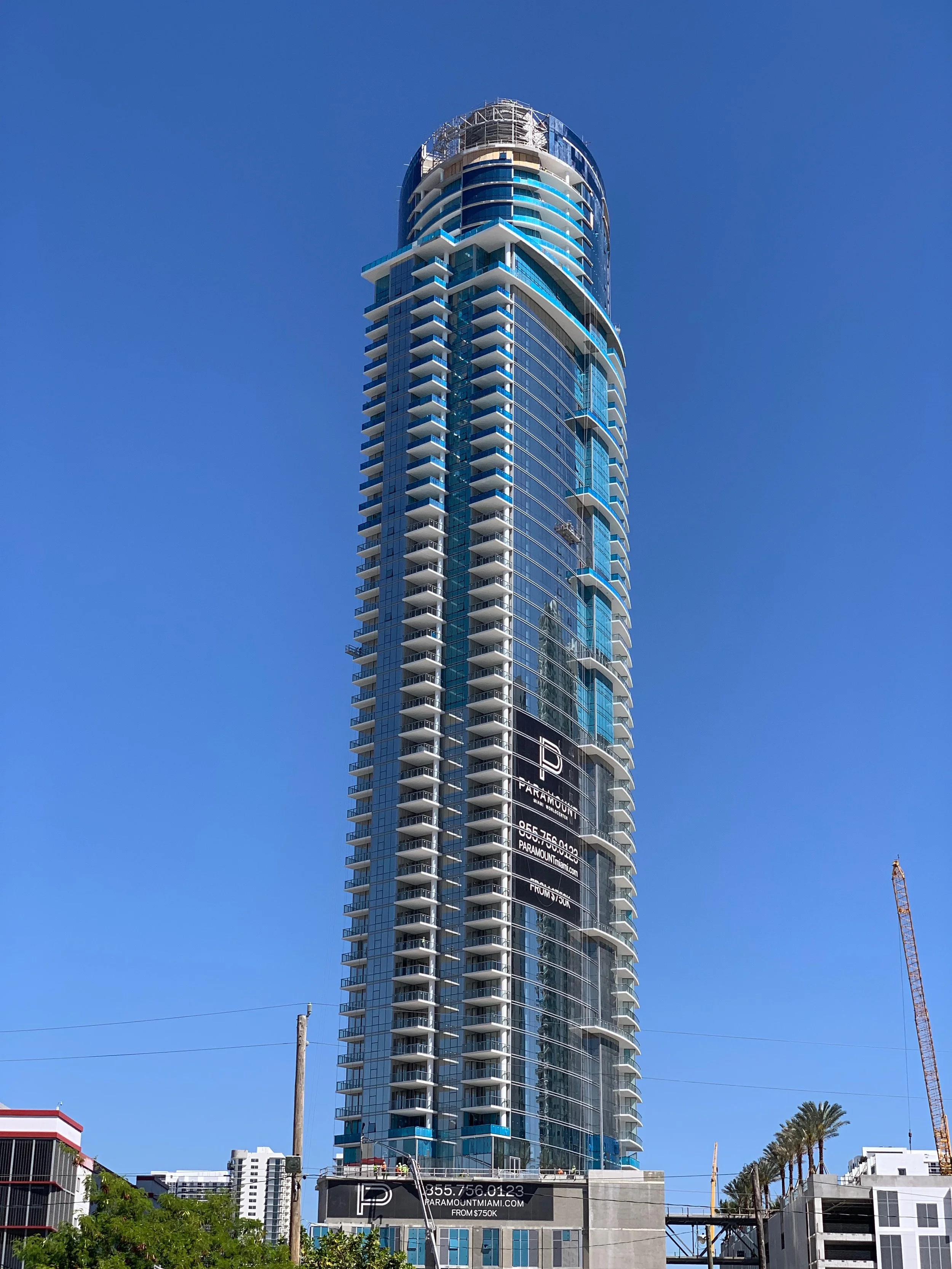 A tall modern skyscraper of Paramount Miami Worldcenter with blue glass windows and multiple balconies, against a clear blue sky. There are palm trees and other buildings at the base.