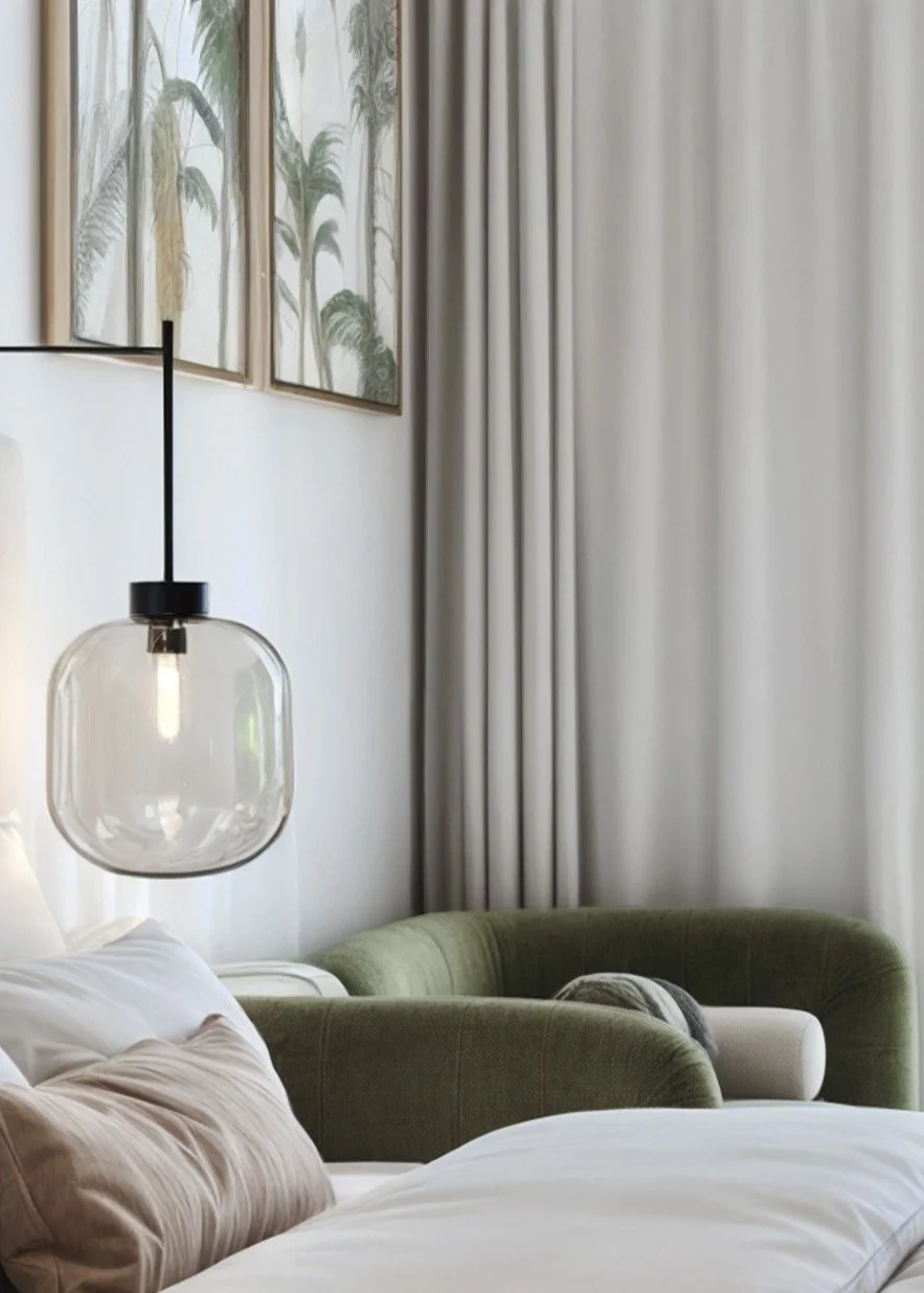 Interior of a bedroom with a bed, green armchair, hanging glass pendant light, framed botanical art on the wall, and cream curtains.
