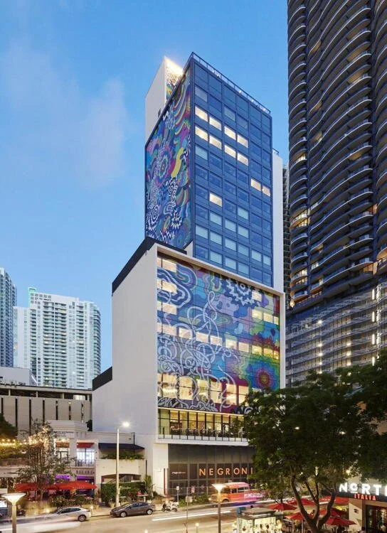 Tall modern building of citizenM Miami Brickell with colorful mural art on its facade, in an urban cityscape at dusk, featuring other high-rise buildings and a busy street below.