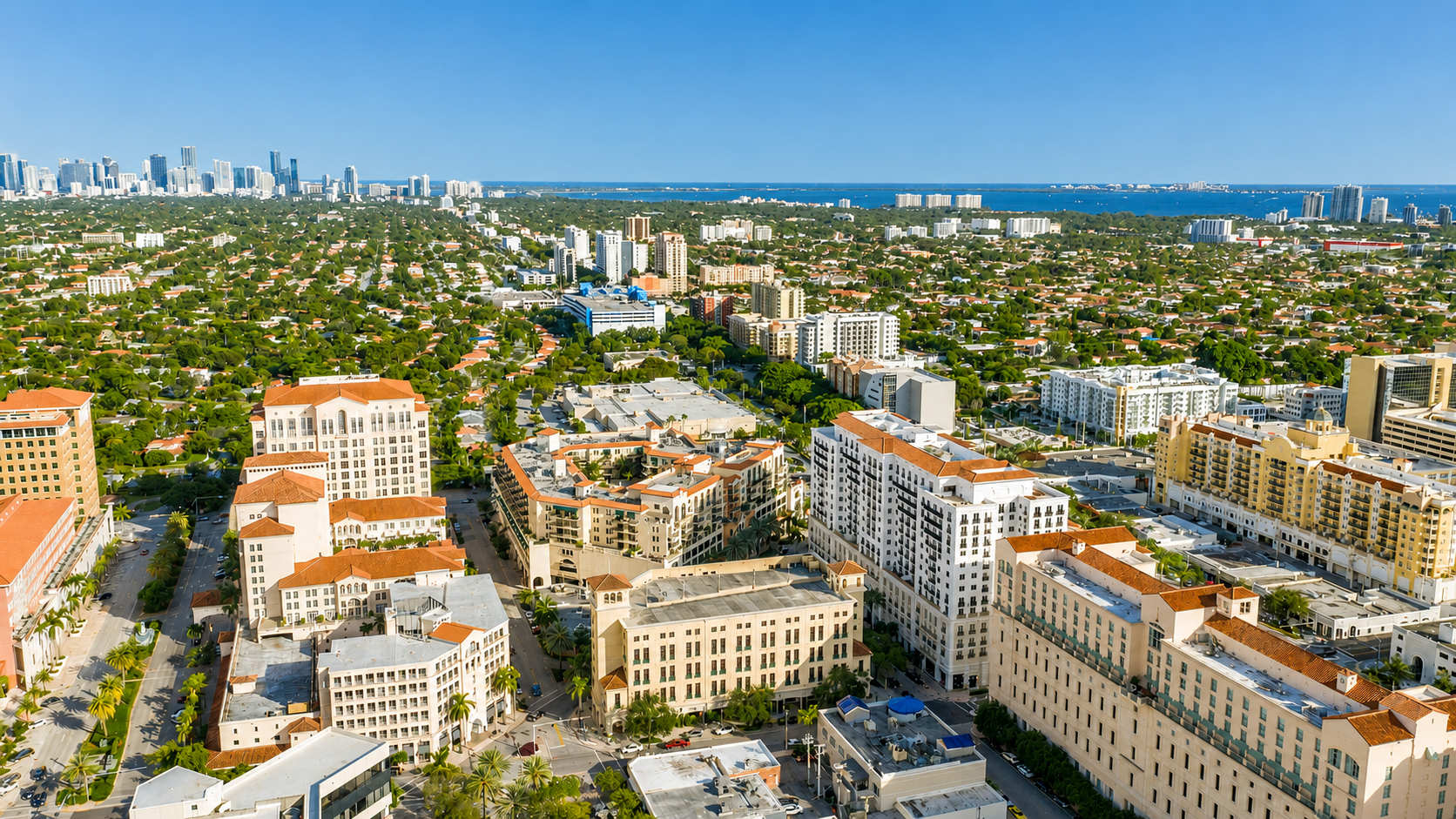 Aerial view of Coral Gables on a sunny day, showing a mix of tall buildings, residential areas, greenery, and a waterfront in the distance.