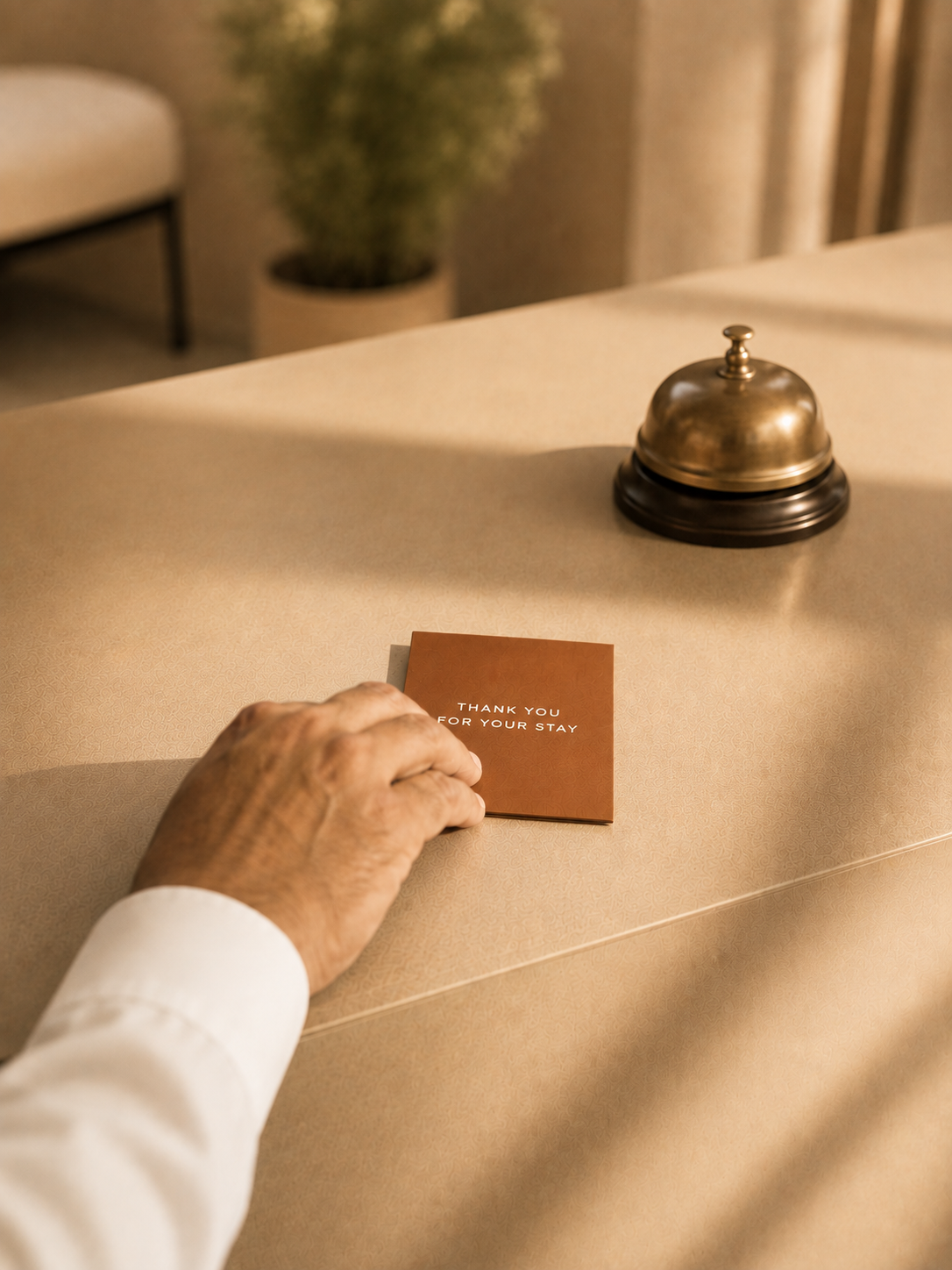Hand placing a thank you card on a hotel reception desk, a bell and potted plant in background.