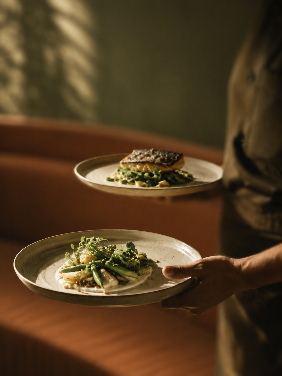 A person serving dishes of gourmet food, including a plate of assorted vegetables and a piece of grilled fish, in a dimly lit setting.