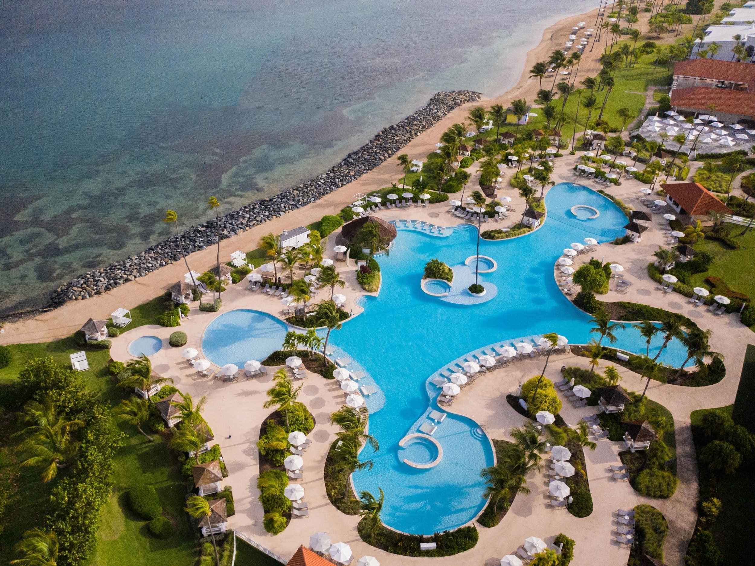 Aerial view of a resort, Hyatt Regency Grand Reserve, featuring a large, winding swimming pool surrounded by white lounge chairs and umbrellas, palm trees, buildings with red roofs, and a sandy beach with calm ocean water.
