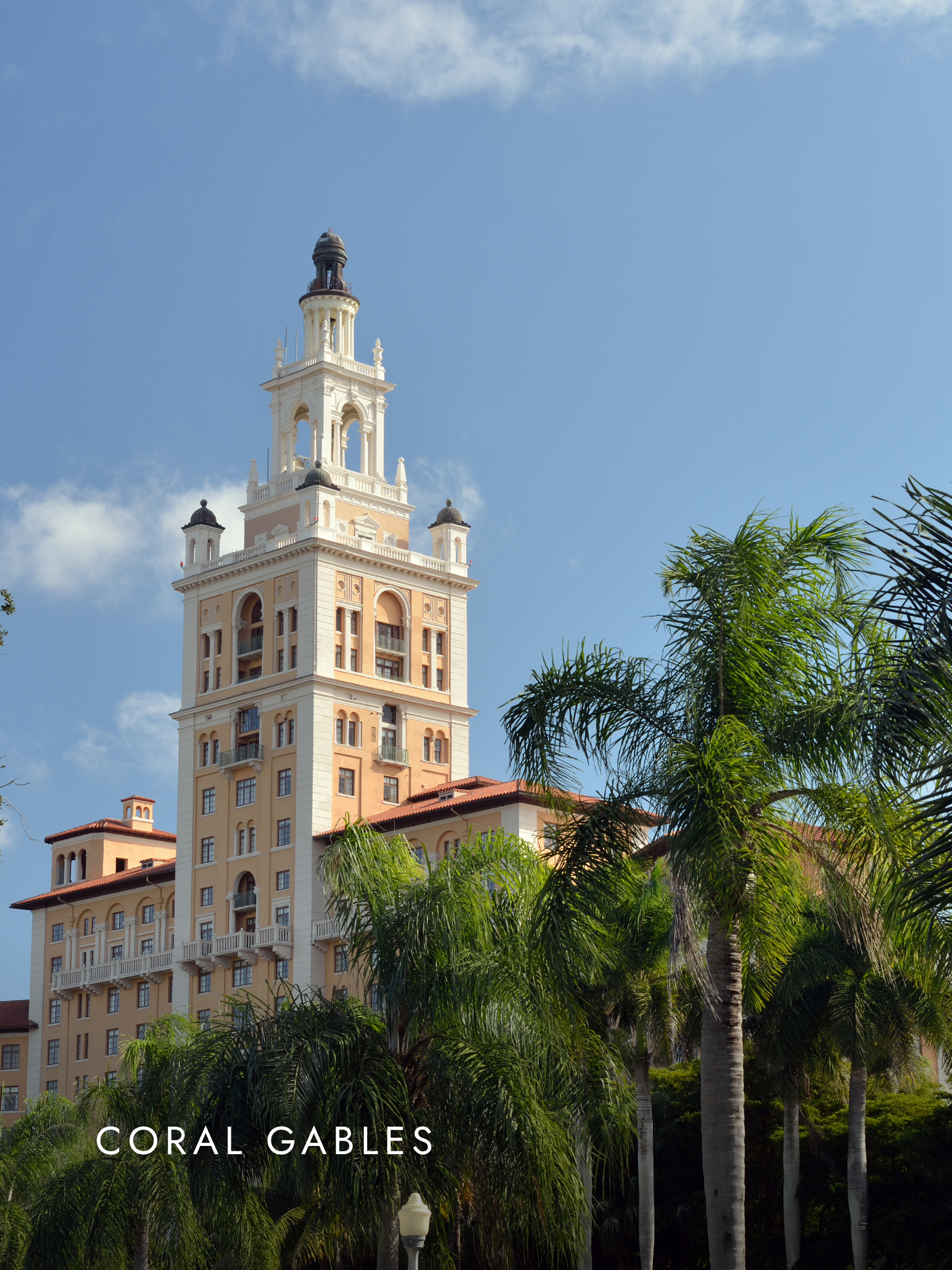 Tall building in Coral Gables with a clock tower surrounded by palm trees under a blue sky.