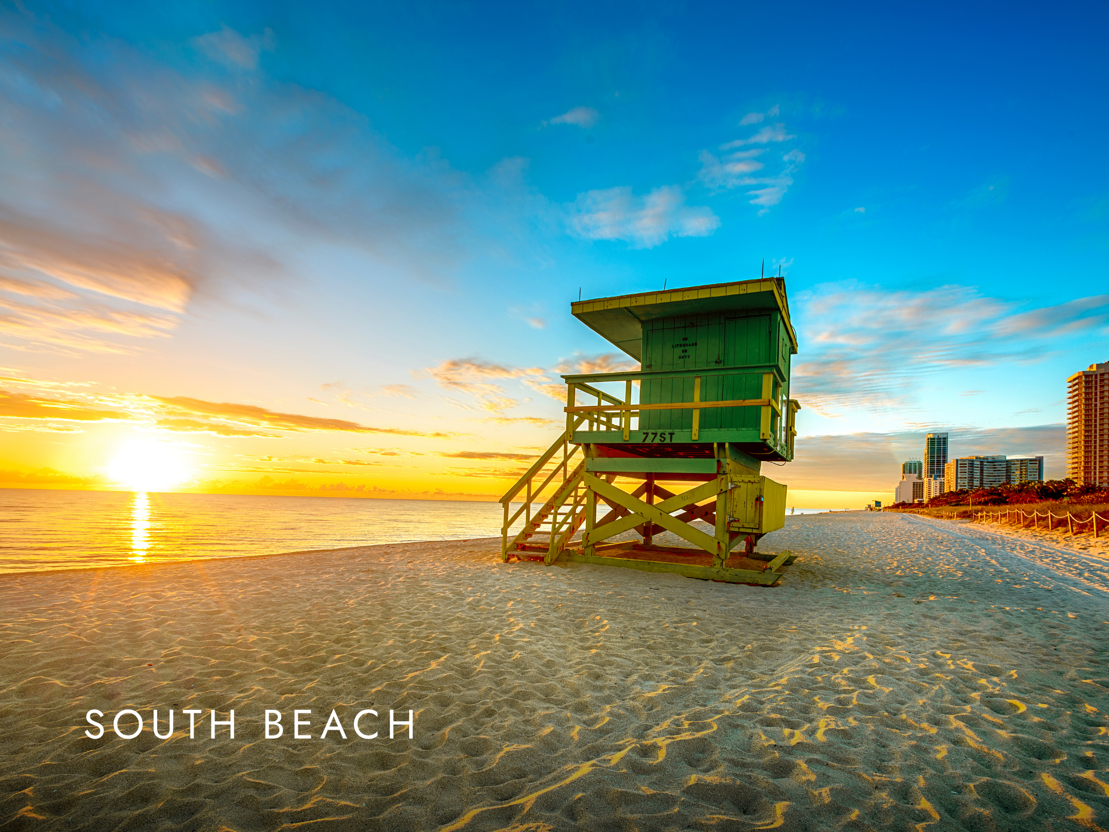A lifeguard tower painted in pale green on the sandy South Beach, with a backdrop of a colorful sunset sky and distant high-rise buildings.