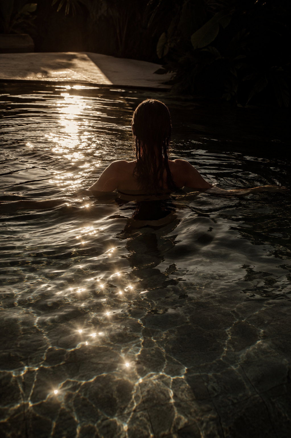 A person with long wet hair swimming in a dark outdoor pool during sunset, with sunlight reflecting off the water.